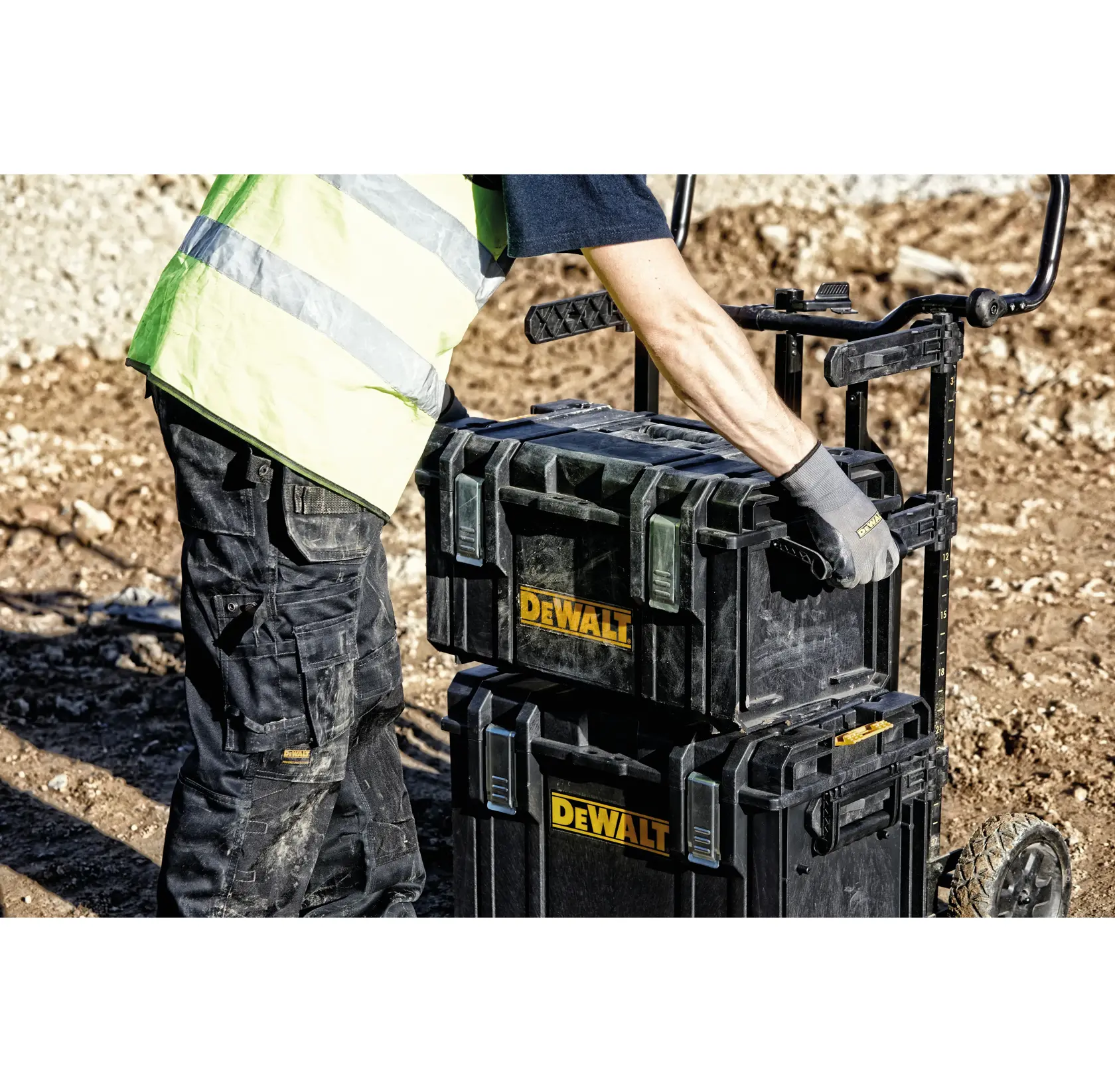 Una persona con chaleco de alta visibilidad y guantes de trabajo mueve cajas de herramientas DEWALT apiladas en un carro en un sitio de construcción.
