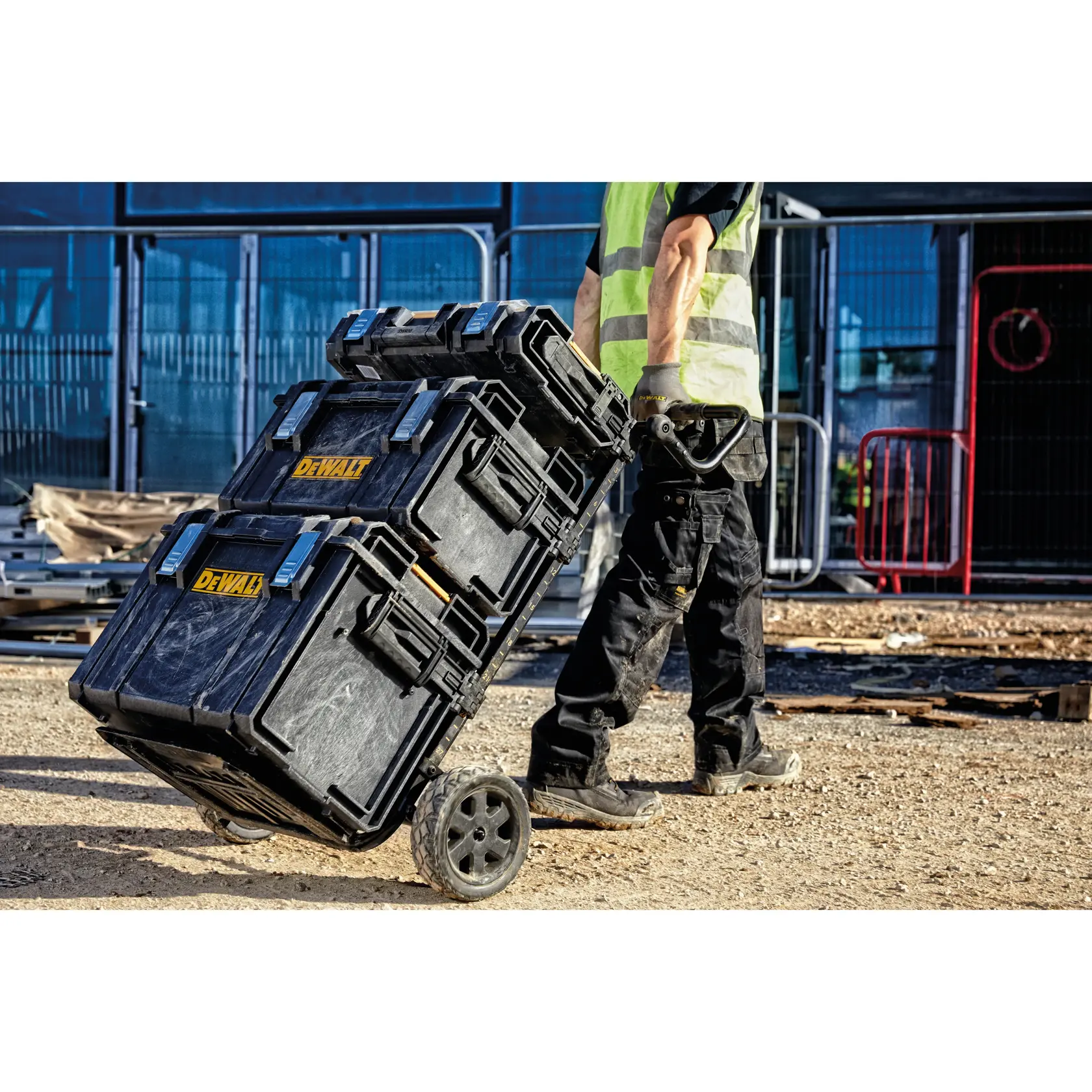 Una persona con ropa de trabajo y chaleco reflectante transporta tres cajas de herramientas negras DEWALT apiladas en un carro con ruedas en una obra de construcción.