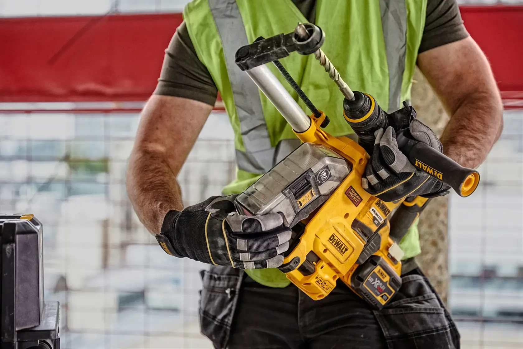 Una persona con chaleco de seguridad verde y guantes sostiene un martillo perforador inalámbrico DEWALT D25304DH con sistema de extracción de polvo integrado en una obra.