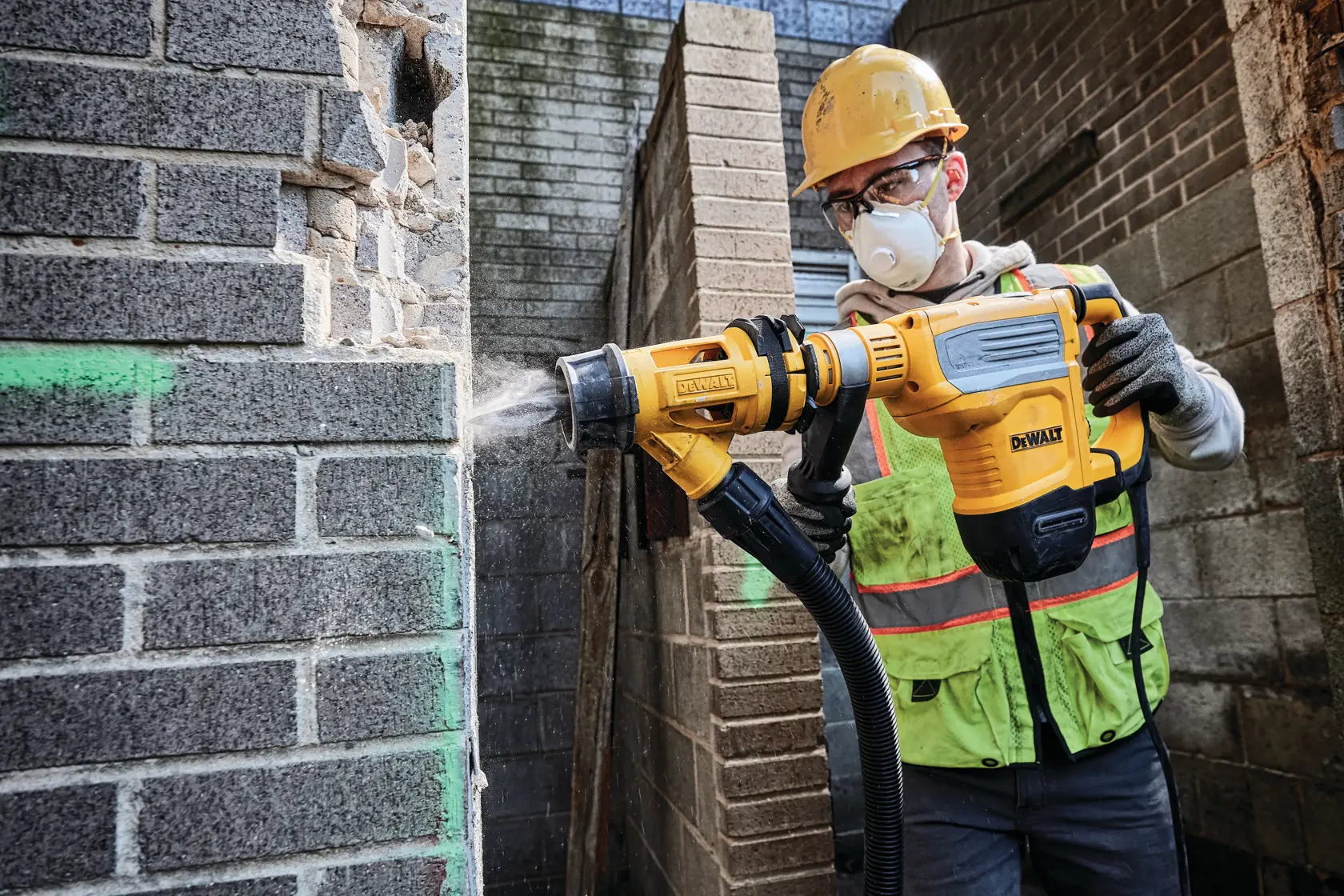 Un trabajador de la construcción con casco amarillo y chaleco de seguridad utiliza un martillo rotatorio DEWALT (modelo D25614K) para romper una pared de ladrillos, mientras el polvo es expulsado por un sistema de extracción de polvo acoplado.