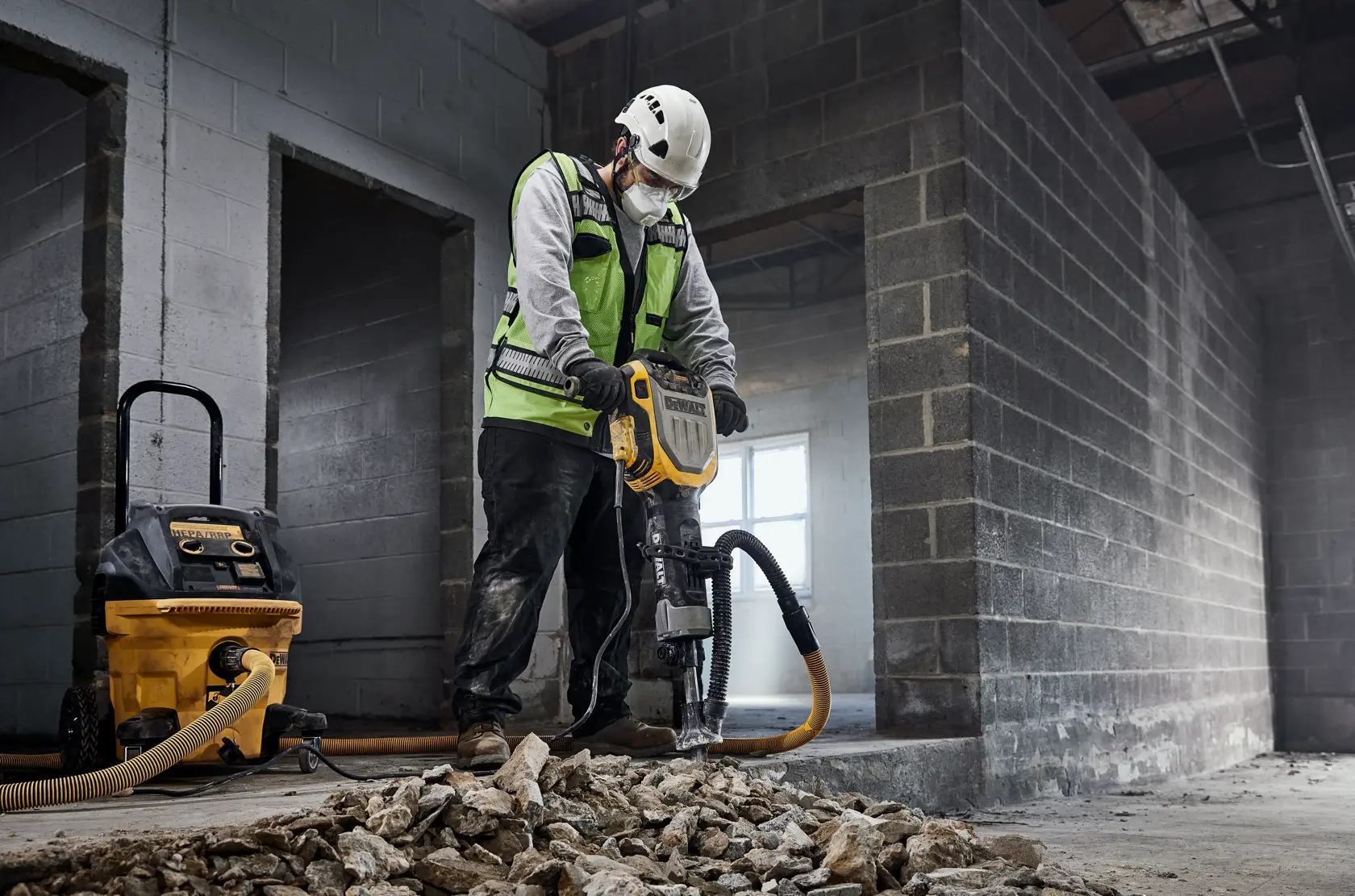 Un travailleur portant un casque de sécurité et une veste haute visibilité utilise un marteau démolisseur DEWALT pour casser du béton dans un environnement industriel. Divers outils et équipements de construction, dont un aspirateur et des outils électriques, sont visibles en arrière-plan.