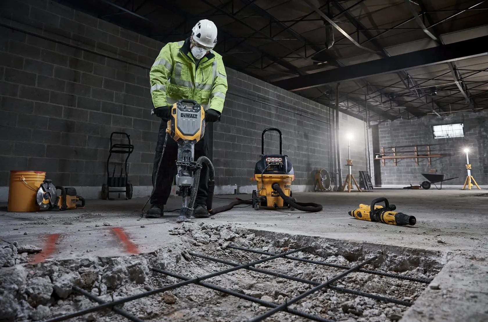 A worker wearing a safety helmet and a high-visibility jacket uses a DEWALT demolition hammer to break up concrete in an industrial setting. Various construction tools and equipment, including a vacuum and power tools, are visible in the background.