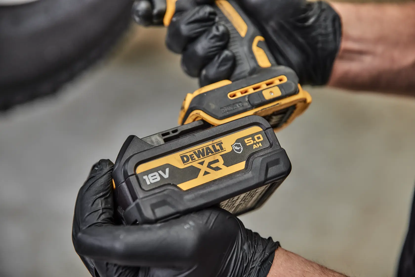 Vista en primer plano de una persona con guantes negros sosteniendo una herramienta eléctrica inalámbrica DEWALT con una batería de litio-ion XR de 18V DCB184G.