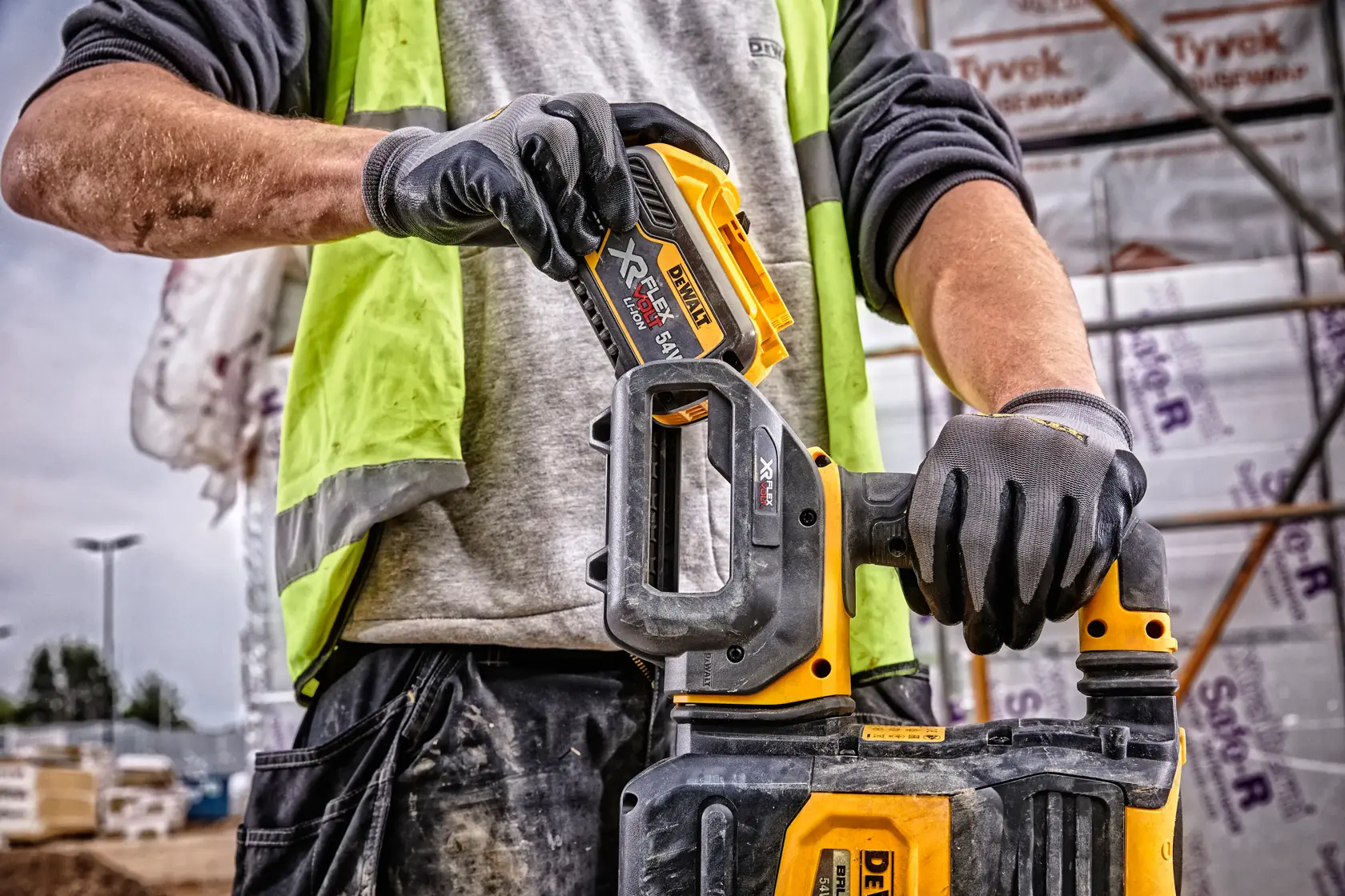 Primer plano de una persona con guantes y chaleco de alta visibilidad, insertando una batería DEWALT DCB548 XR FLEXVOLT en una herramienta eléctrica compatible DEWALT en una obra de construcción.