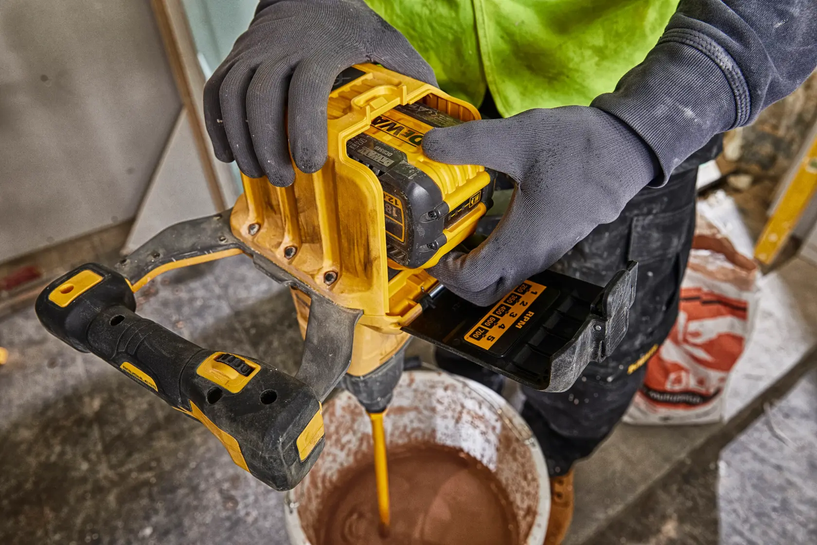 Una persona con guantes utiliza una mezcladora inalámbrica amarilla DEWALT con una batería DEWALT DCB548 para mezclar material en un balde en un sitio de construcción.