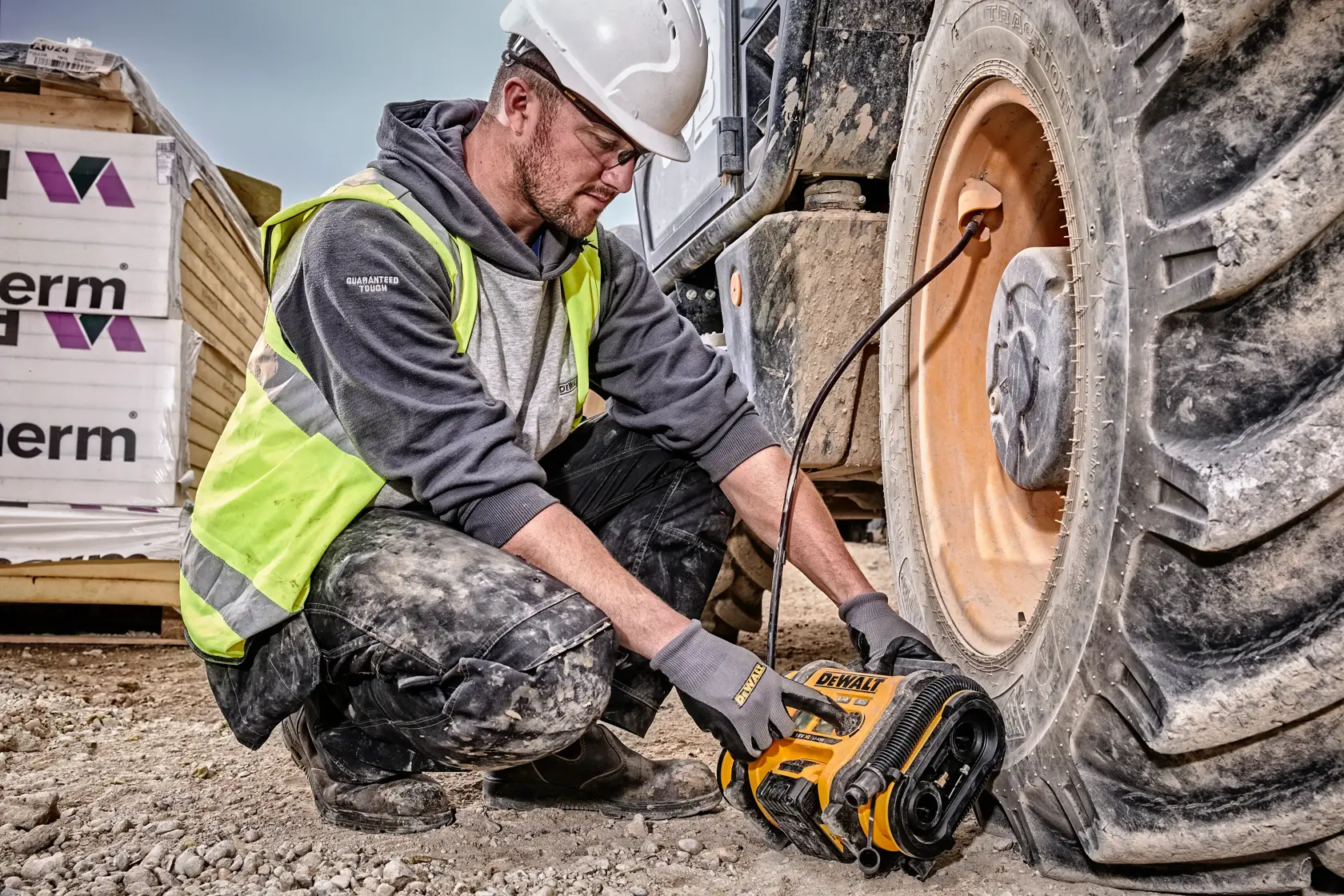 Una persona con casco de seguridad y chaleco reflectante está utilizando un compresor de aire inalámbrico DEWALT DCC018N para inflar el neumático de un vehículo de construcción grande en una obra.