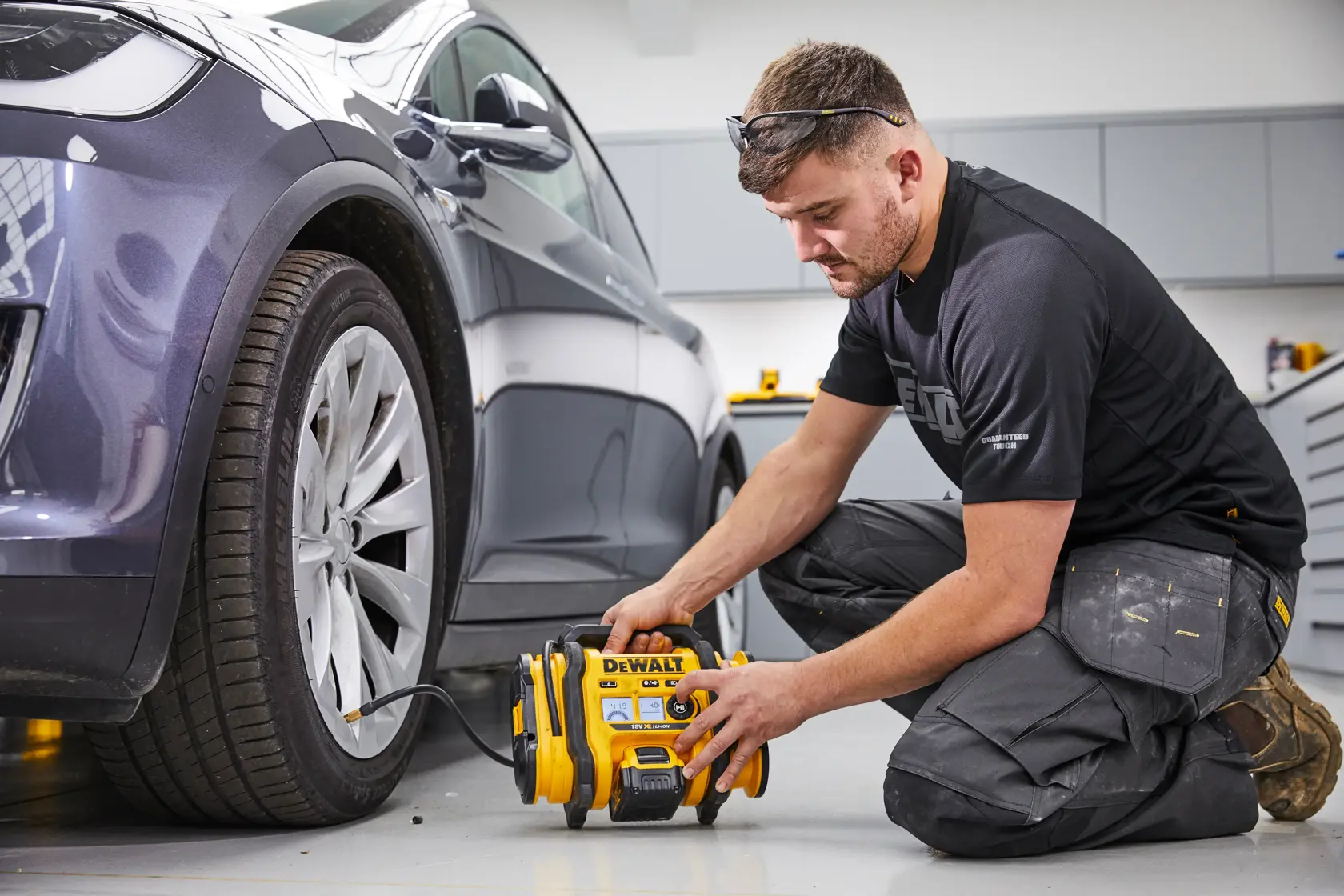 Una persona arrodillada en el suelo utiliza un compresor de aire portátil DEWALT DCC018N para inflar el neumático de un coche estacionado en un garaje o taller. El rostro de la persona está difuminado por privacidad.
