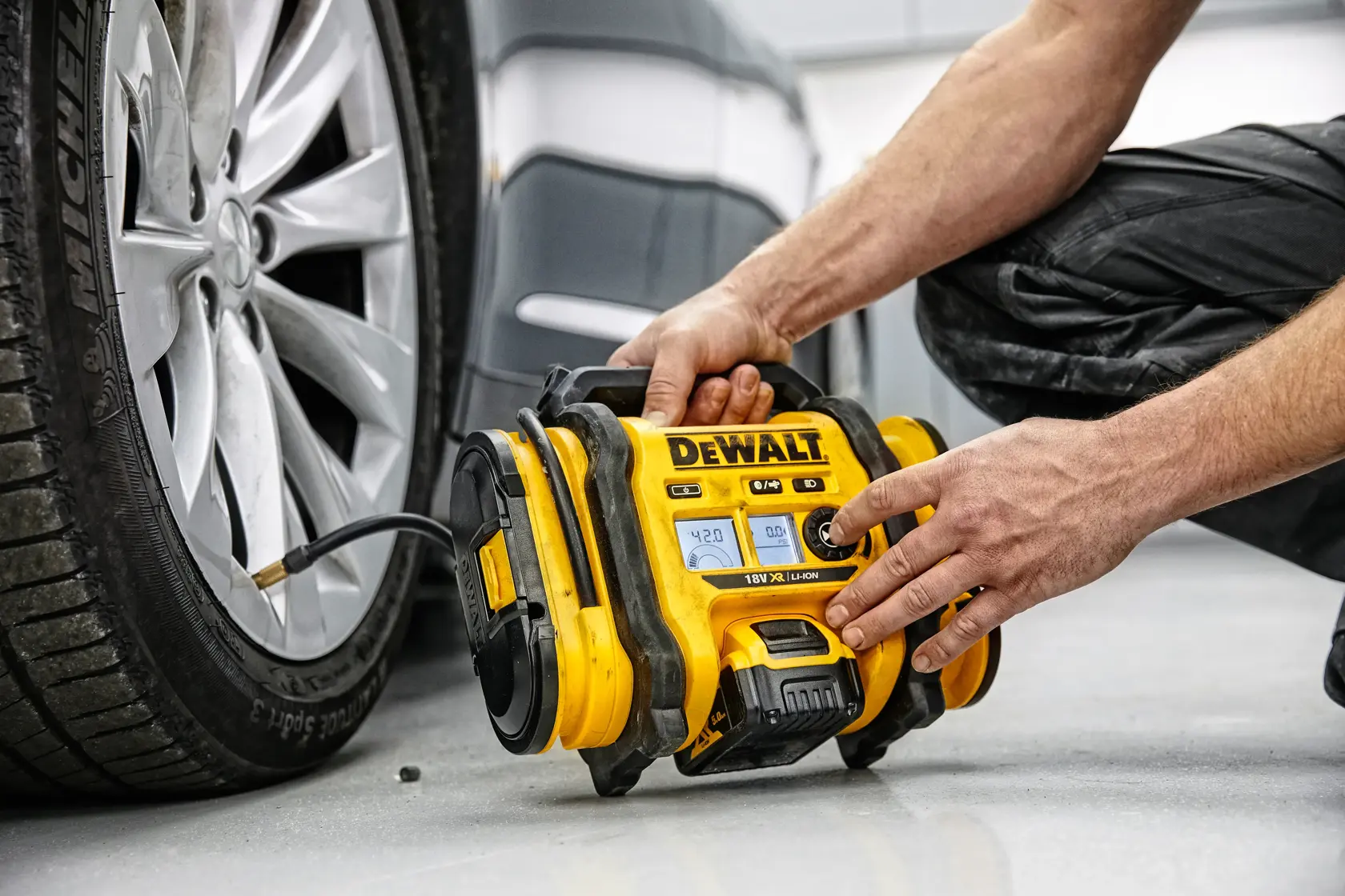 Una persona utiliza un compresor de aire portátil amarillo DEWALT para inflar la llanta de un coche. El dispositivo está conectado a la válvula del neumático y colocado en el suelo junto al vehículo.