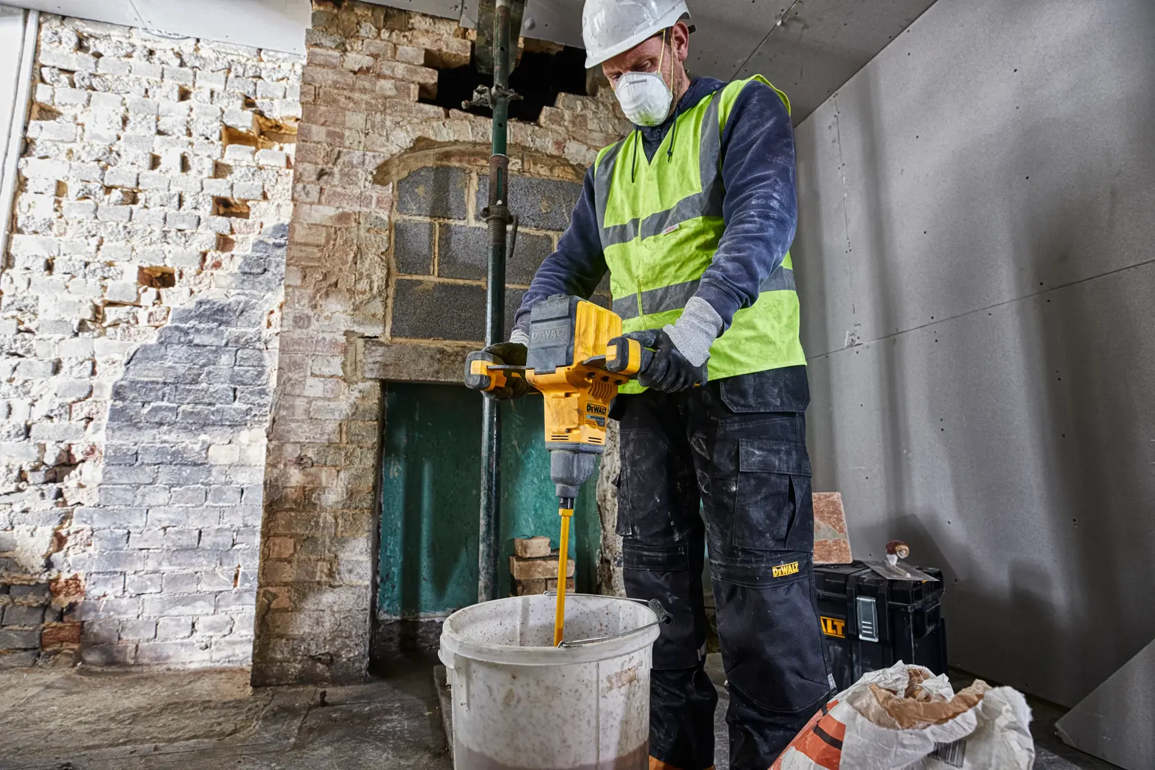 Una persona con chaleco de seguridad y guantes utiliza un mezclador inalámbrico DEWALT DCD240X2 para remover el contenido de un gran balde en un sitio de construcción con paredes de ladrillo y paneles de yeso.