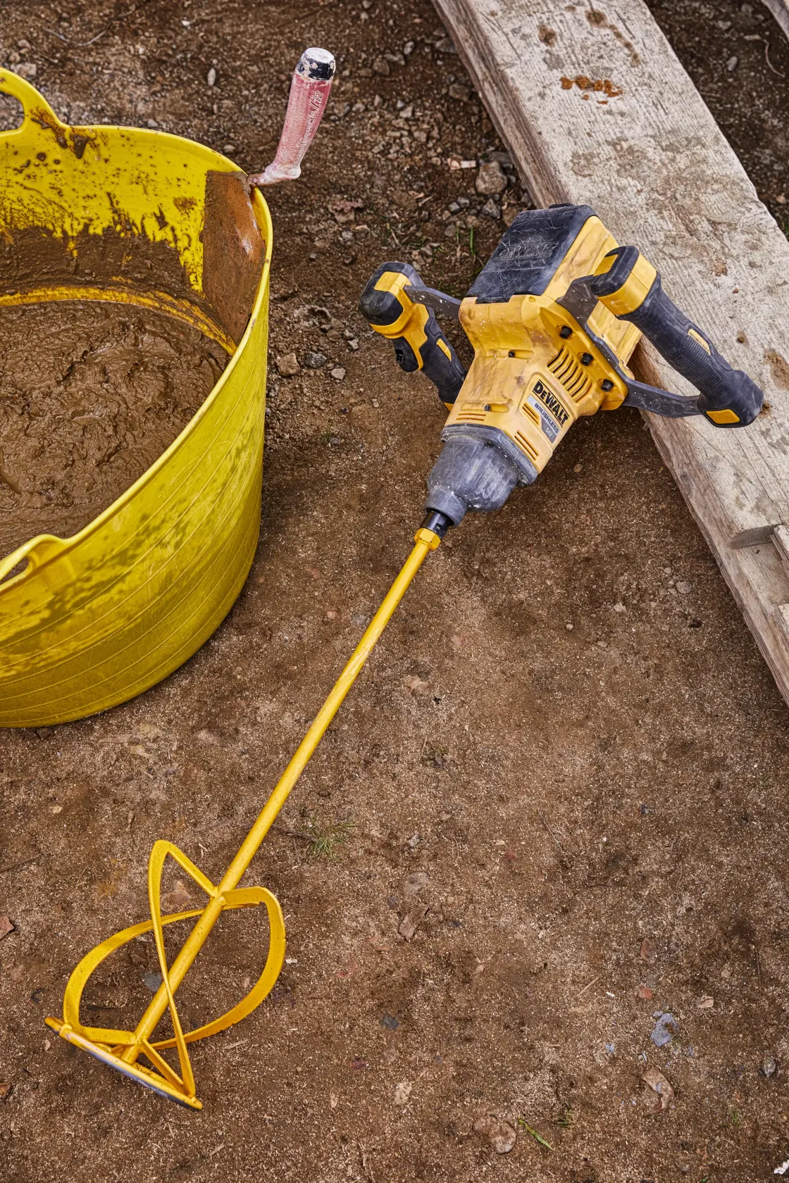 Un mezclador inalámbrico DEWALT con una larga varilla mezcladora amarilla está sobre el suelo de una obra junto a un cubo amarillo que contiene material mezclado y una paleta.
