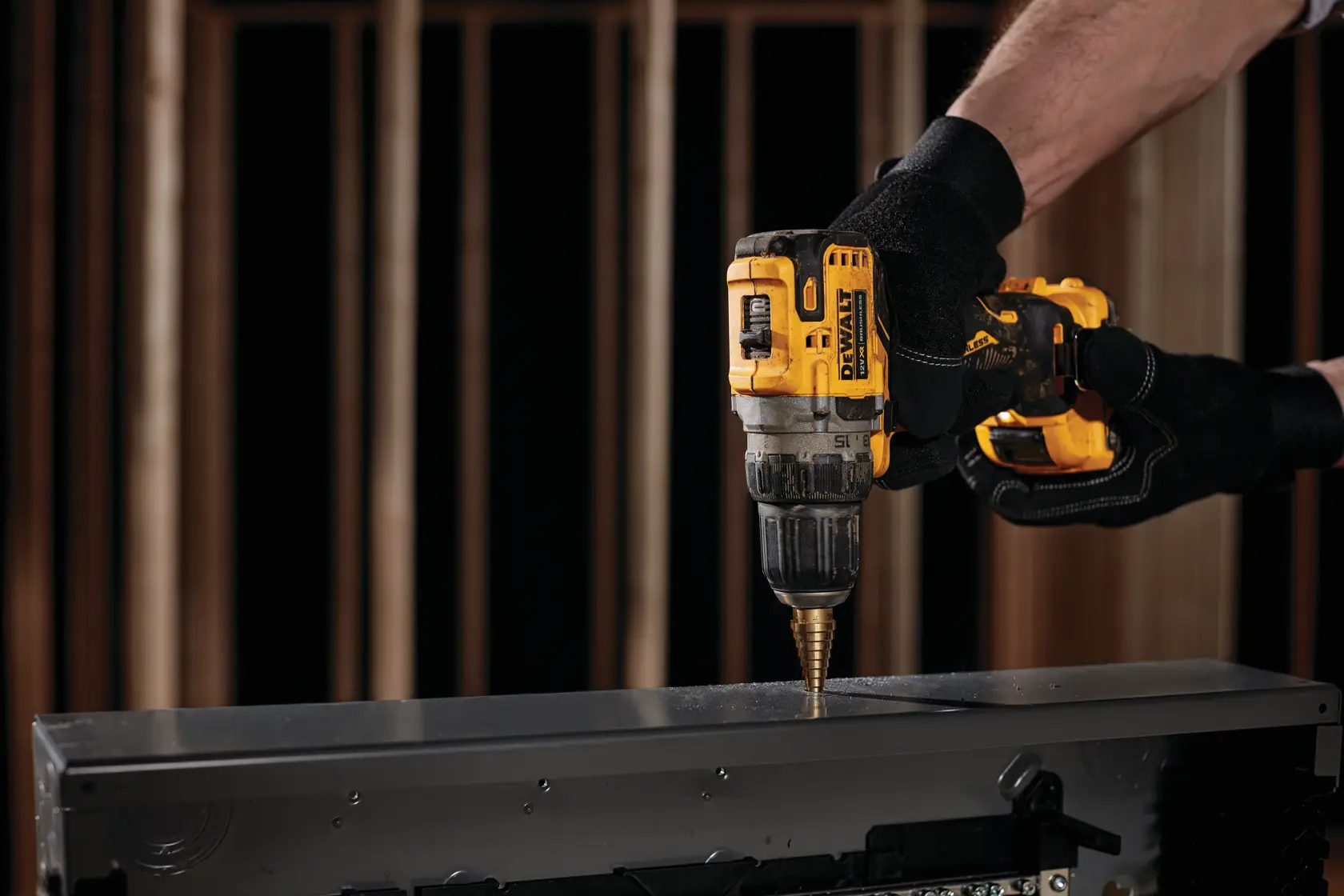 Una persona con guantes negros utiliza un taladro inalámbrico amarillo DEWALT para hacer un agujero en una superficie metálica.