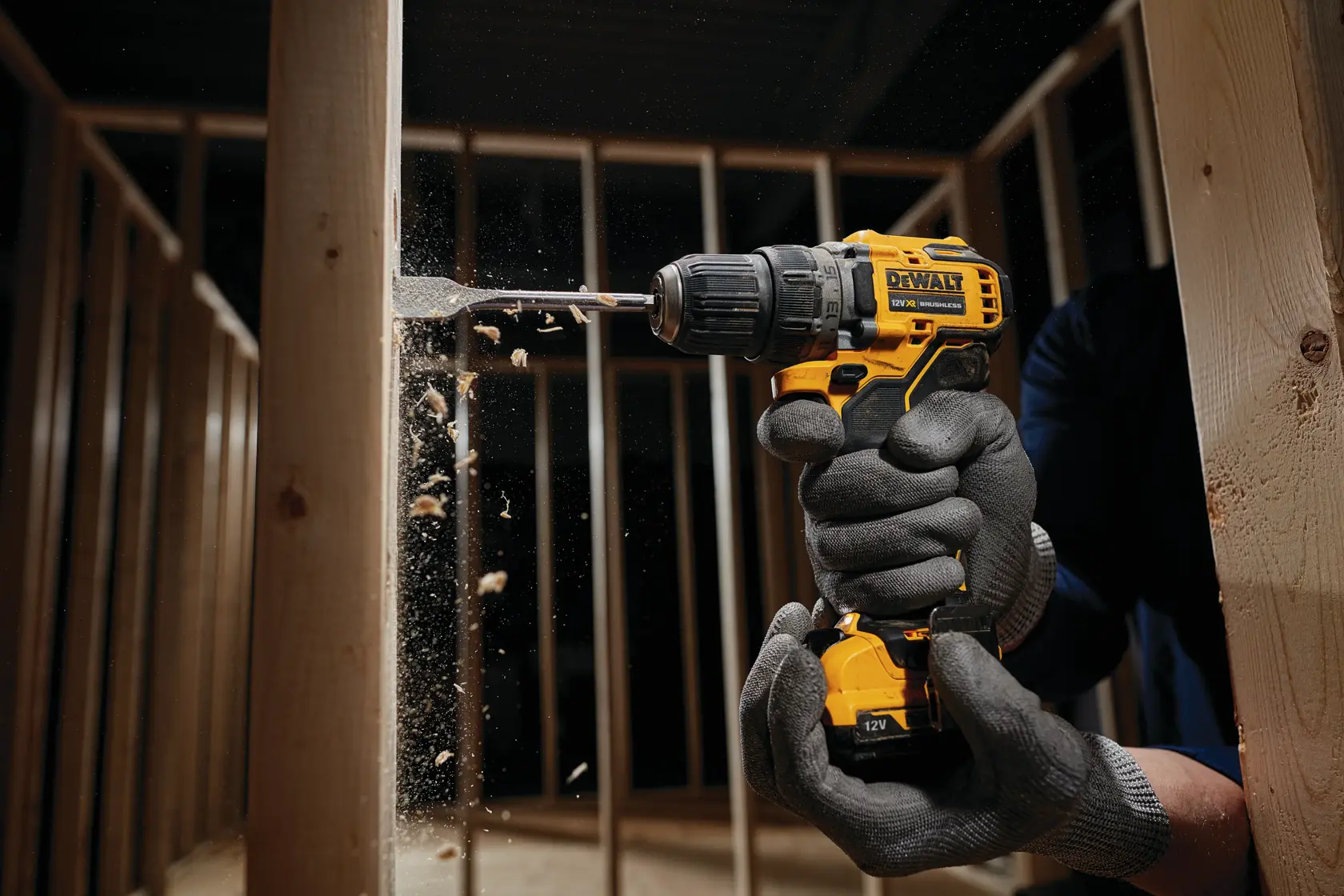 Una persona con guantes protectores utiliza un taladro inalámbrico amarillo DEWALT para perforar una viga de madera dentro de una estructura enmarcada. Se ven virutas de madera volando durante el proceso de perforación.