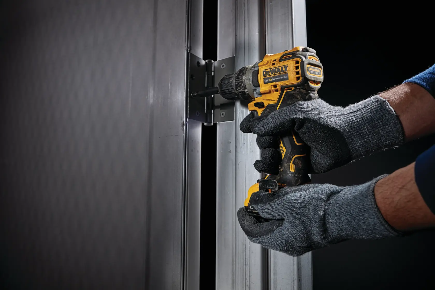Una persona con guantes grises utiliza un taladro inalámbrico amarillo y negro DEWALT para asegurar una bisagra en un marco de puerta de metal.