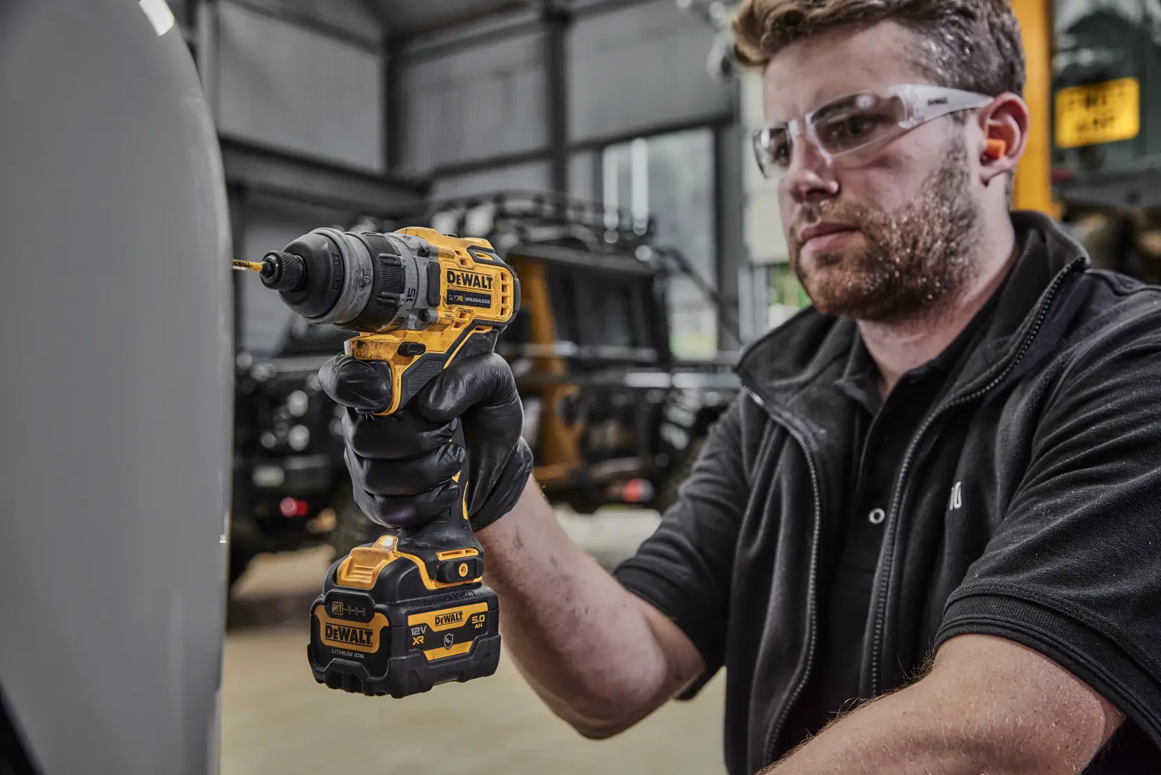 Una persona con guantes negros está utilizando un taladro inalámbrico DEWALT amarillo y negro con una batería adjunta en interiores. La cara de la persona está difuminada por privacidad.