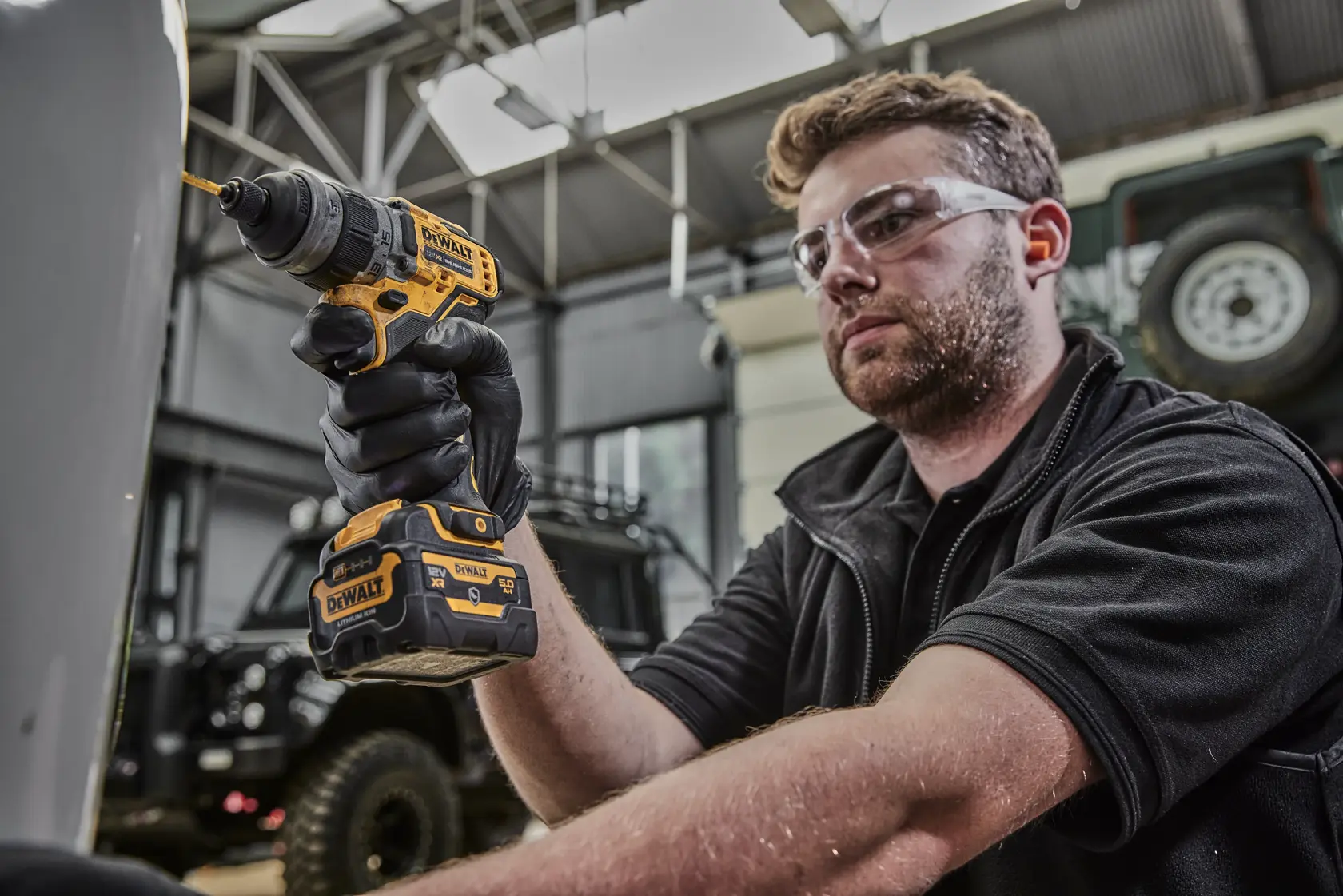 Una persona con el rostro difuminado está usando un taladro inalámbrico DEWALT DCD703NT amarillo y negro en un taller o garaje. La persona lleva guantes negros.