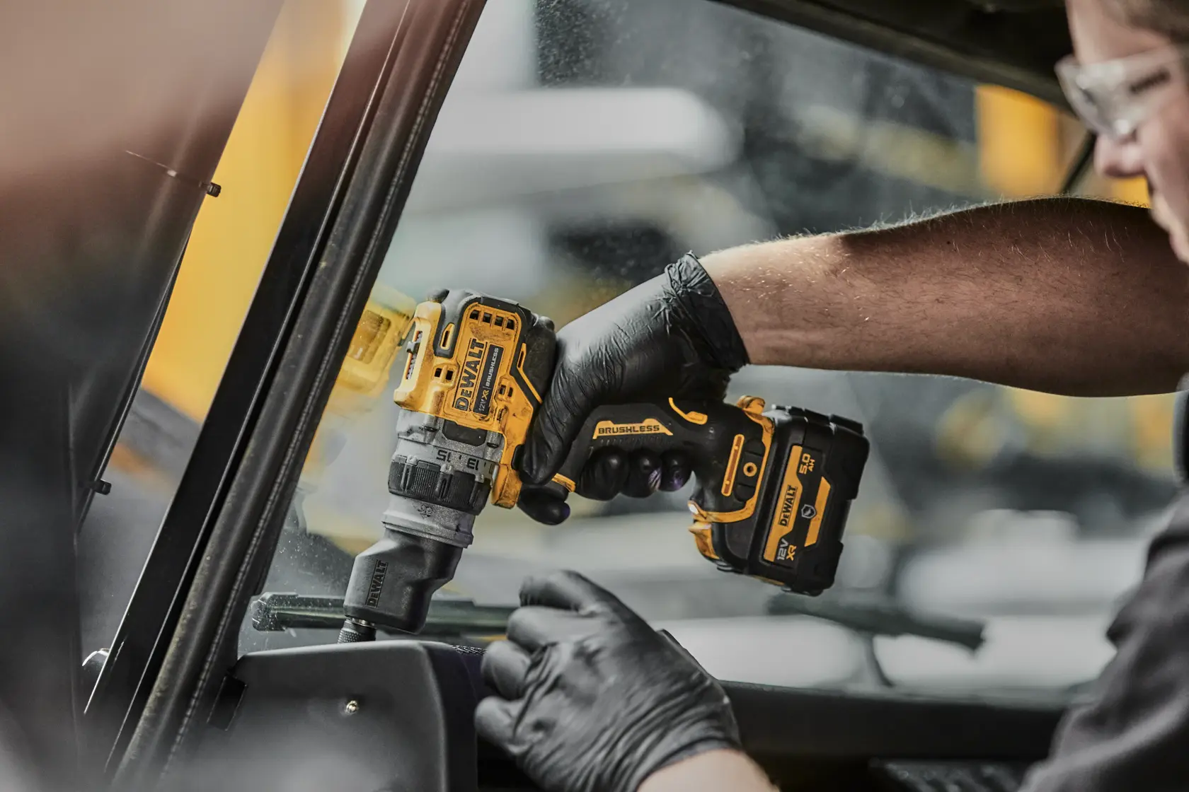 Una persona con guantes negros utiliza un taladro inalámbrico amarillo y negro DEWALT con motor sin escobillas para trabajar dentro de un vehículo cerca de una ventana.