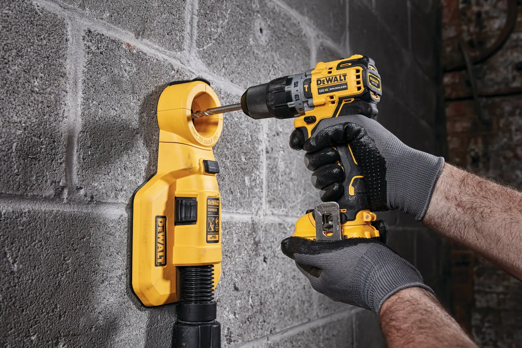 Una persona con guantes grises utiliza un taladro inalámbrico amarillo DEWALT para perforar una pared de concreto, con un extractor de polvo amarillo acoplado para recoger los residuos.
