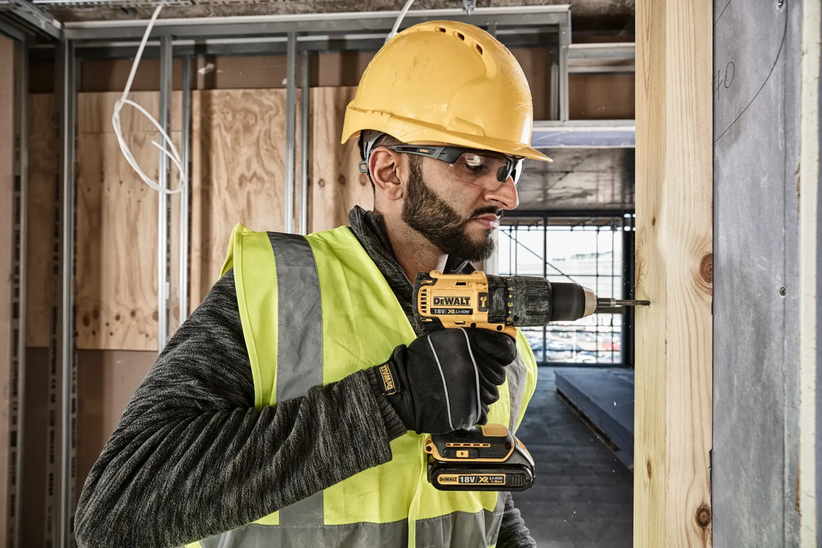 Una persona con casco de seguridad amarillo y chaleco reflectante utiliza un taladro inalámbrico DEWALT DCD778S2T para perforar una tabla de madera en un sitio de construcción. El rostro de la persona está difuminado por privacidad.