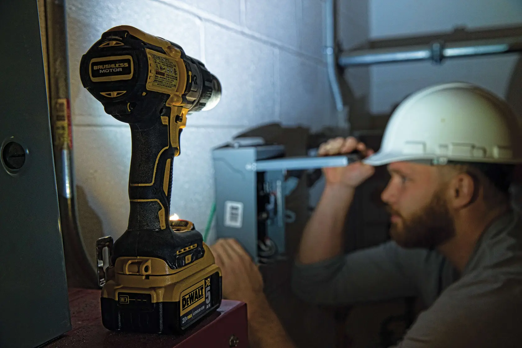 Una persona con chaqueta de seguridad reflectante y casco blanco utiliza un taladro inalámbrico DEWALT amarillo y negro para trabajar en una pared de concreto en un entorno industrial.