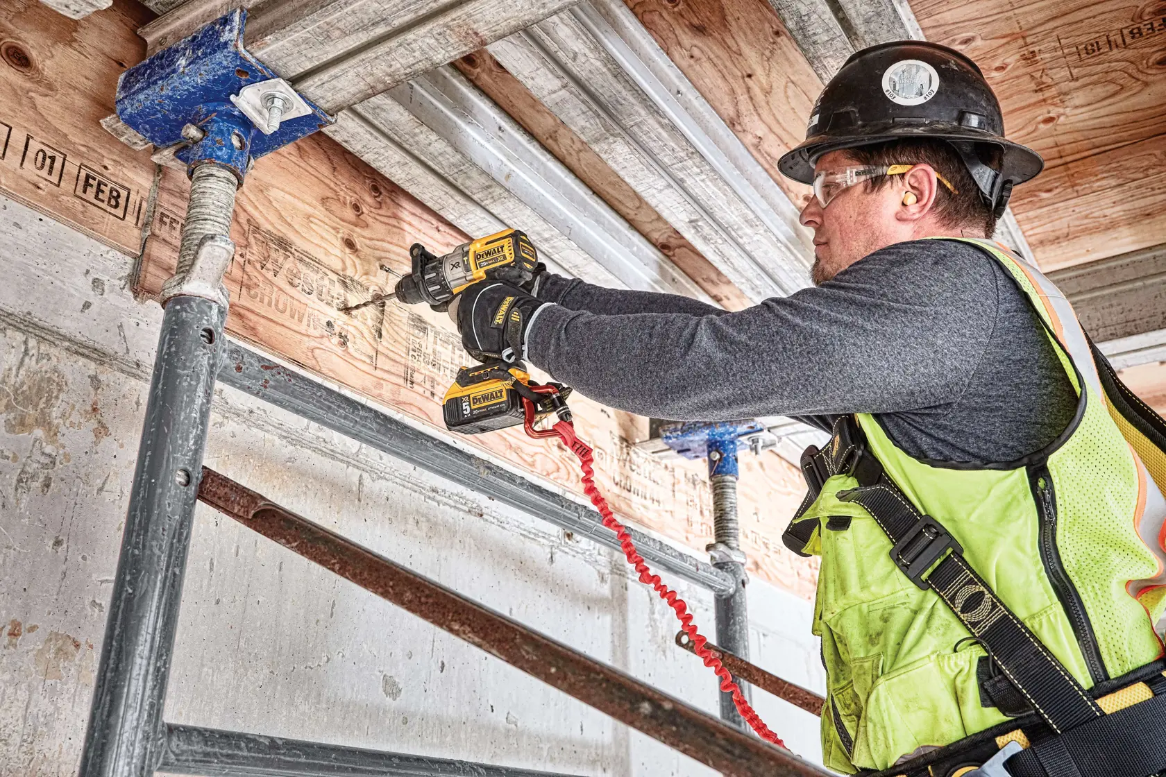 Una persona utiliza un taladro eléctrico DEWALT DCD996P2 para perforar una viga de madera en un sitio de construcción. Hay aserrín en el suelo y la persona lleva botas de trabajo y pantalones vaqueros.
