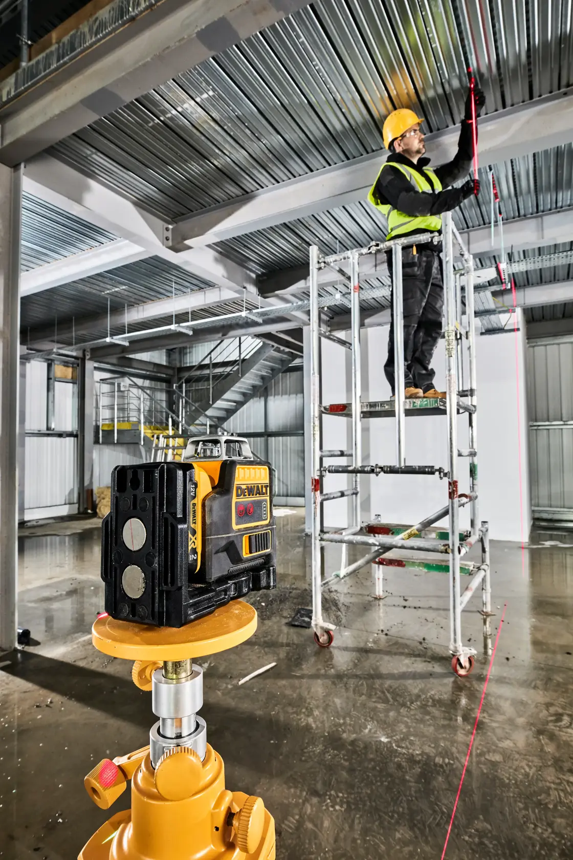 Un nivel láser DEWALT está colocado sobre un trípode en primer plano en un sitio de construcción industrial. Al fondo, un trabajador con casco amarillo y chaleco reflectante está de pie en un andamio, marcando el techo metálico siguiendo la línea láser roja proyectada por el dispositivo. El área incluye vigas metálicas, escaleras y materiales de construcción.