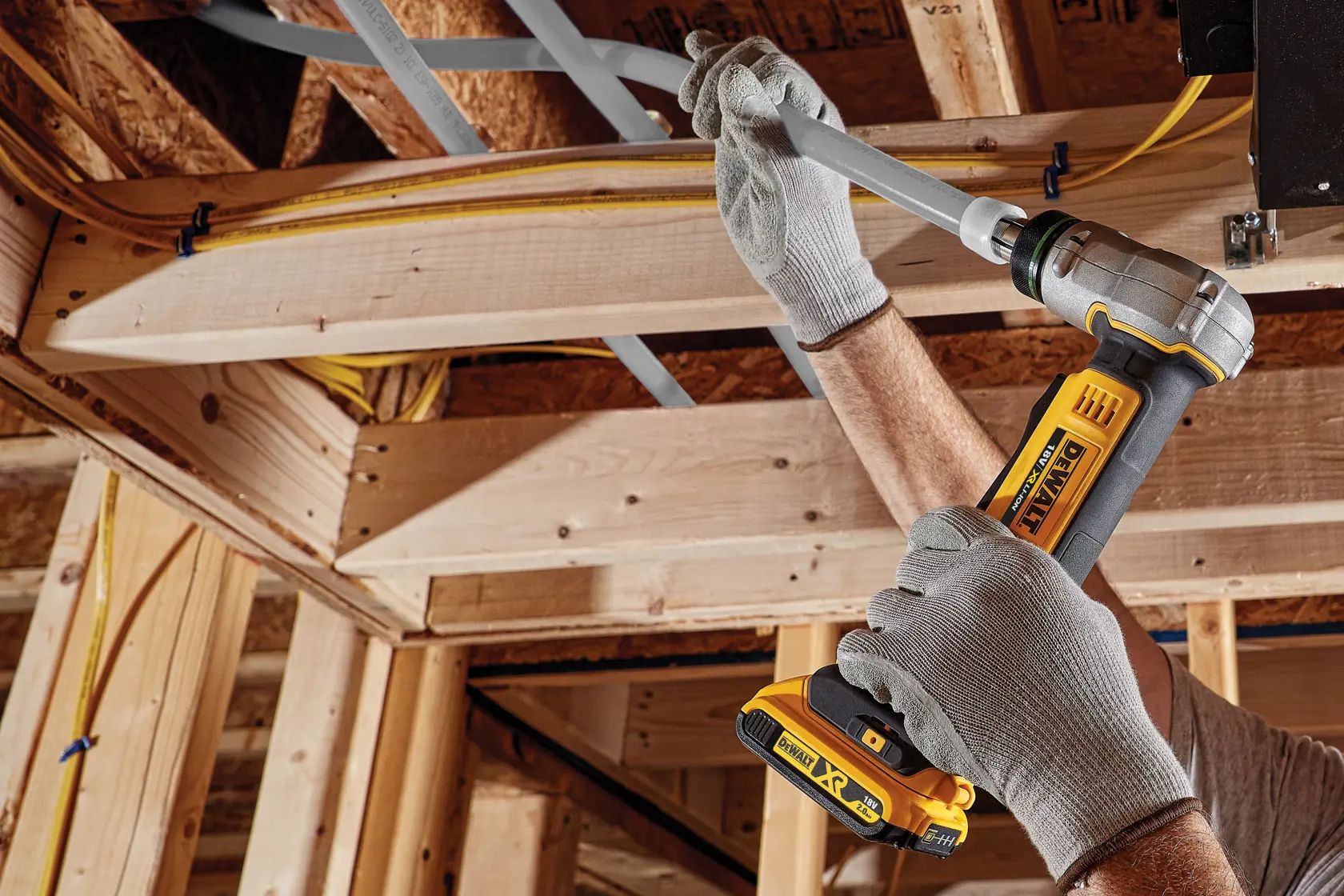 Una persona con guantes de trabajo grises utiliza una herramienta de crimpar tubos inalámbrica DEWALT para asegurar un tubo de plástico gris a una estructura de madera durante la construcción.