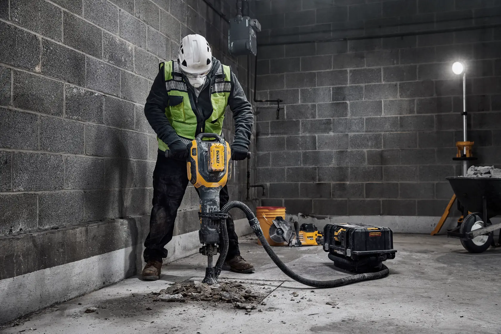 A construction worker wearing a safety helmet and reflective vest uses a DEWALT demolition tool to break concrete in an industrial setting. Nearby are a pipe, a tool case, a dolly, and a standing work light.