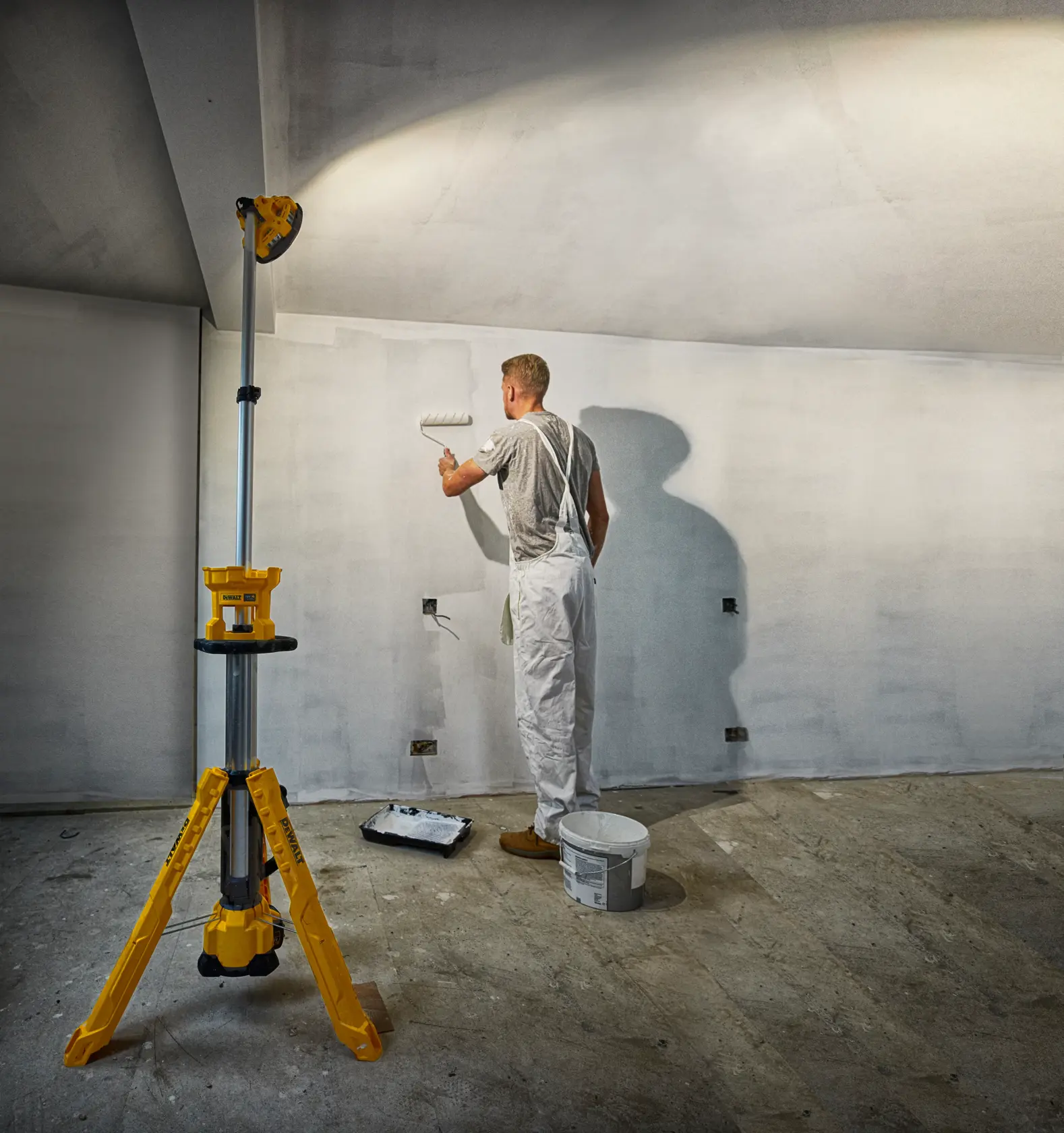 Un pintor usa un rodillo sobre una pared, iluminado por una luz de trabajo DEWALT amarilla de trípode.