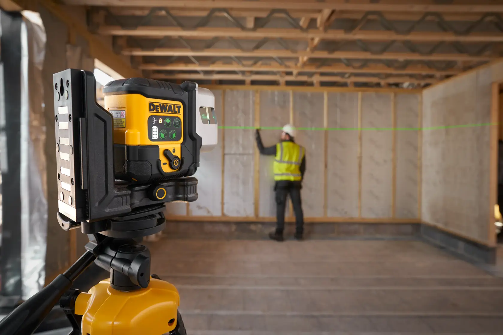 Nivel láser DEWALT en trípode proyecta línea verde, trabajador marca alineación en pared.