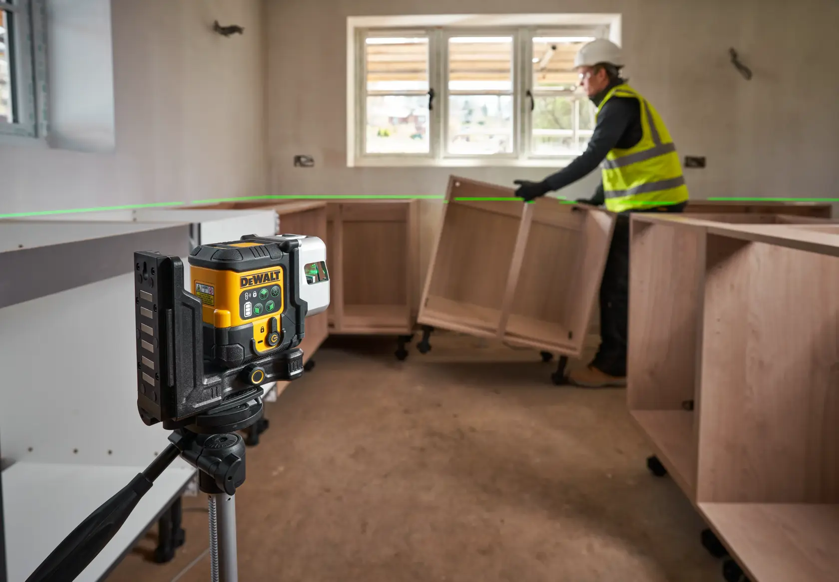 Nivel láser DEWALT en trípode proyecta línea verde en habitación con instalación de muebles.