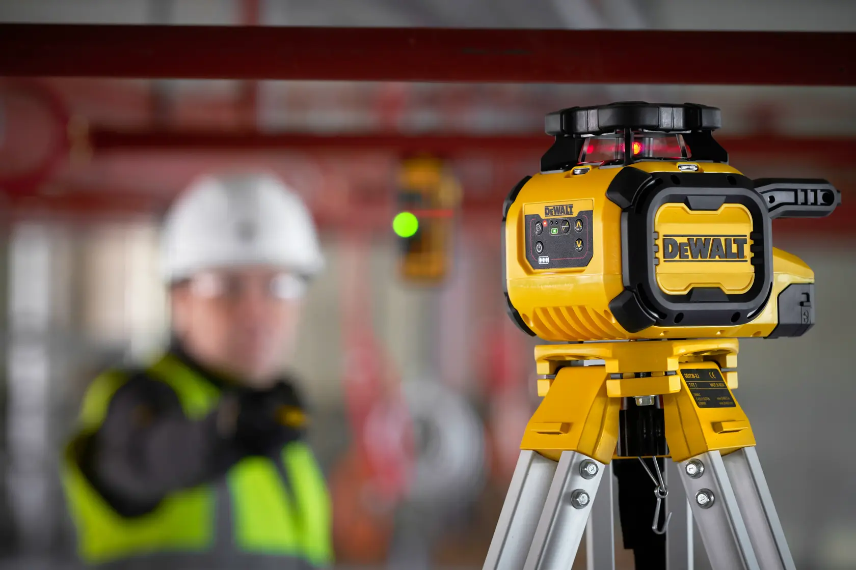 Láser DEWALT en trípode, DCLE15211R y DCLE15211R-QW SKUs, obra, trabajador desenfocado.