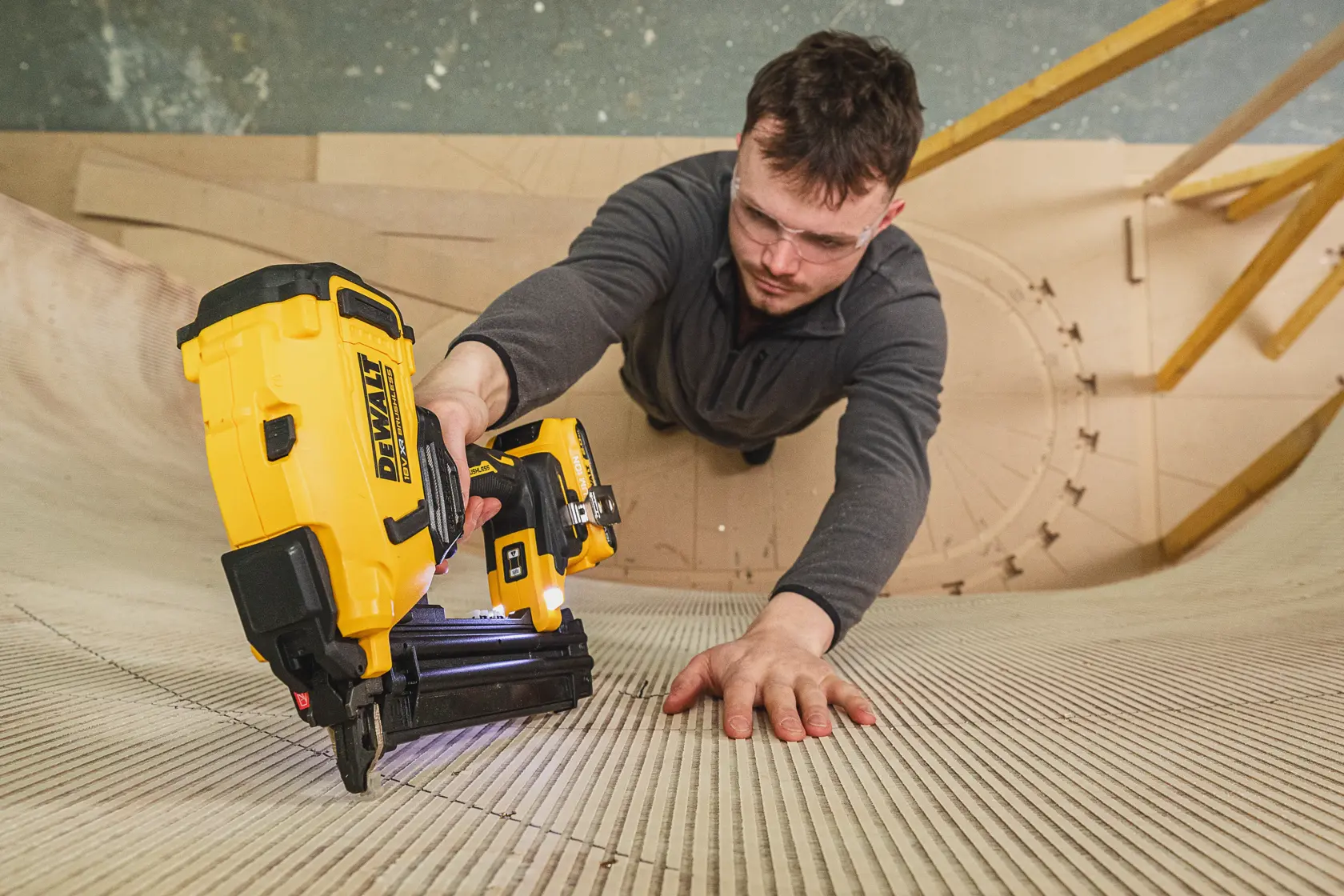 Persona usando una clavadora DEWALT en superficie de madera curvada, lentes de seguridad, ropa gris.