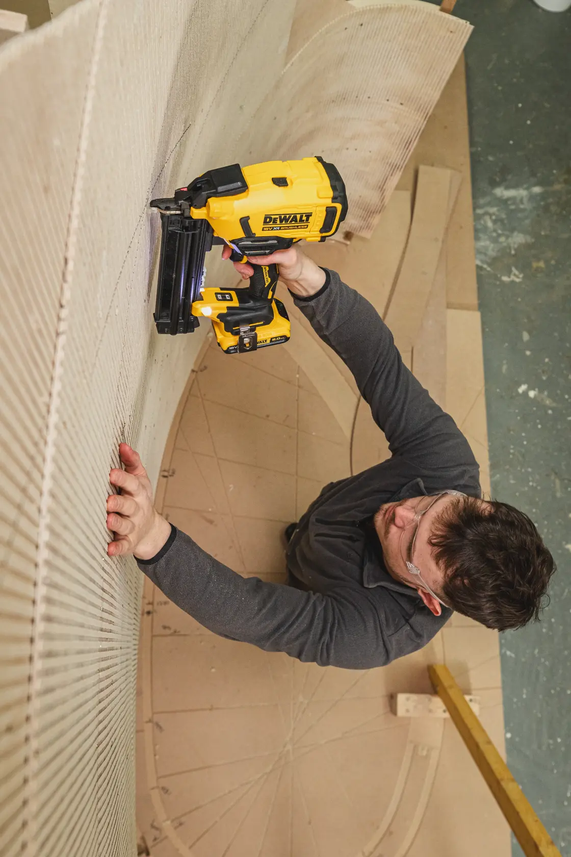 Persona usando clavadora DEWALT DCN210SN-XJ en madera curva en taller, con gafas de seguridad y ropa oscura.