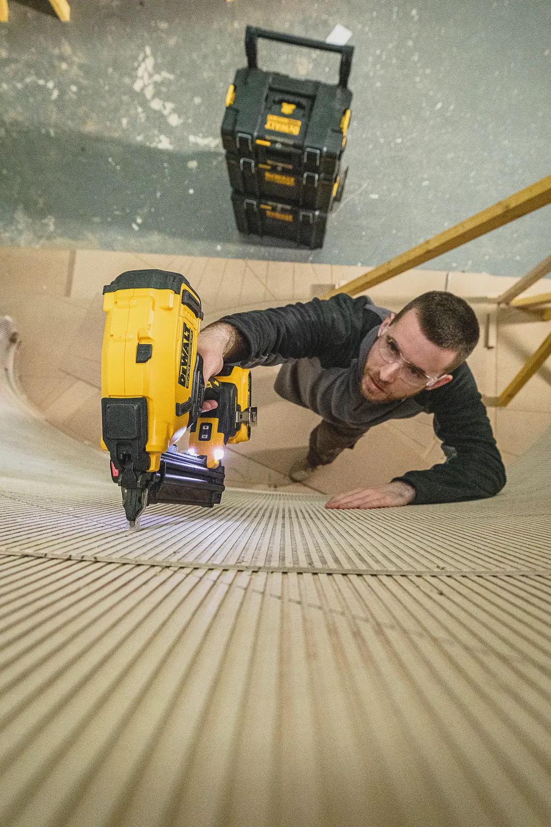 Persona utiliza una clavadora DEWALT en madera, con cajas DEWALT visibles al fondo.
