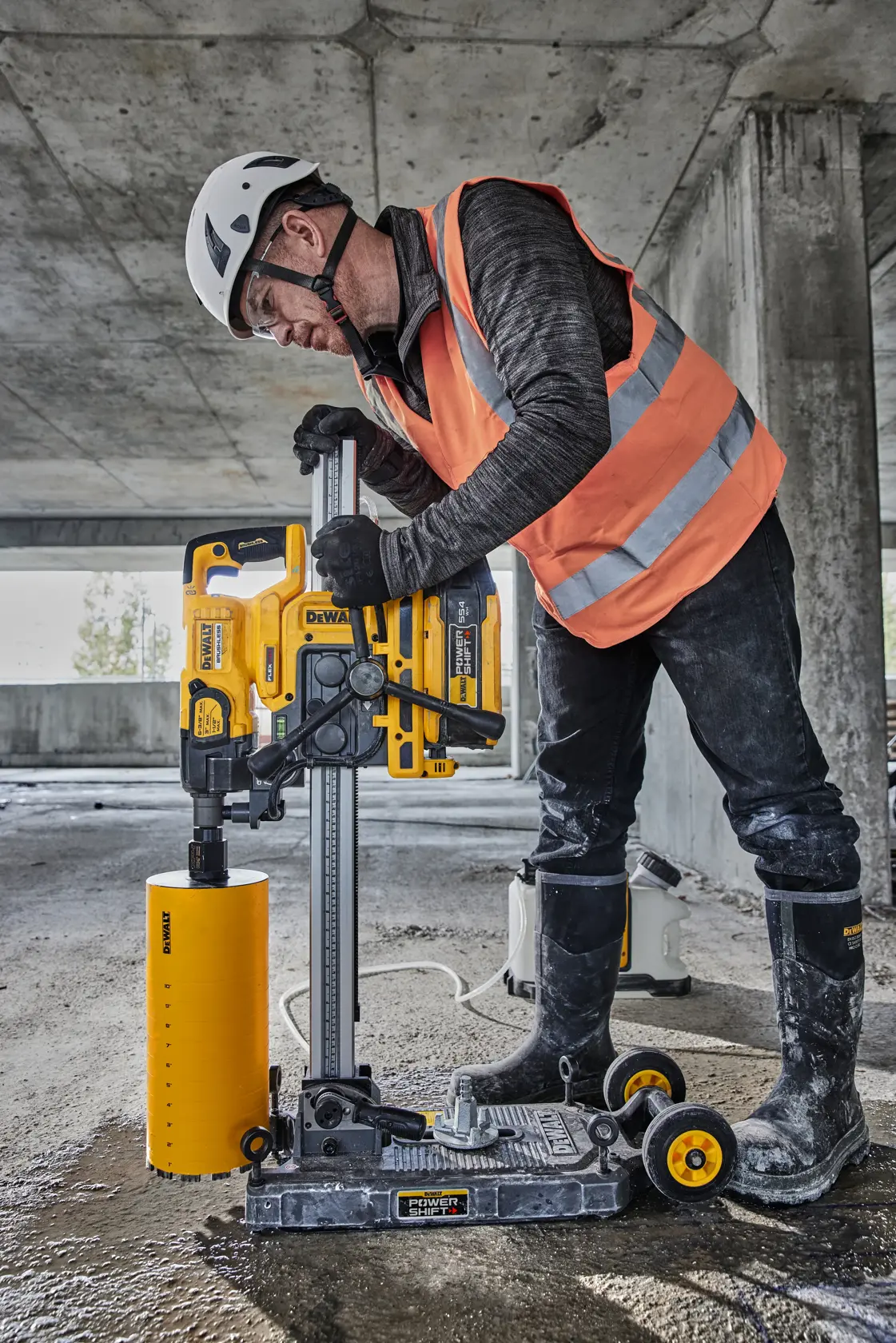 Trabajador de construcción con casco de seguridad y chaleco naranja de alta visibilidad sosteniendo una herramienta DEWALT frente a un camión de obra en un sitio de construcción. La cara del trabajador está desenfocada para proteger su privacidad.