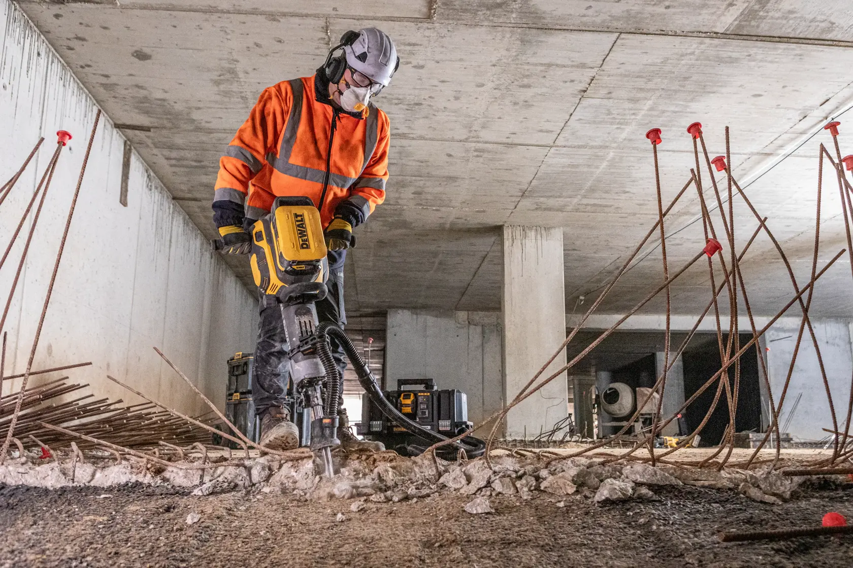 Trabajador usa rompedor DEWALT con equipo de seguridad, rodeado de varillas y escombros.