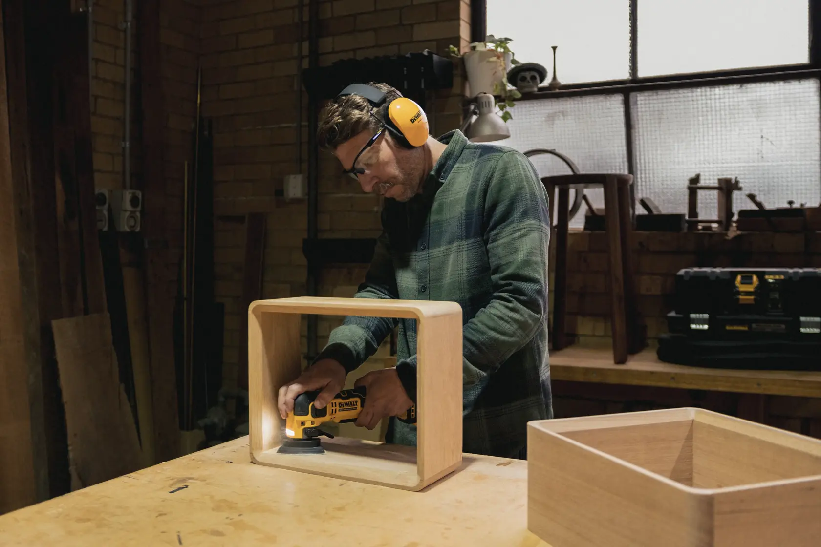 Persona lija caja de madera con herramienta DEWALT en taller, usando equipo de seguridad.