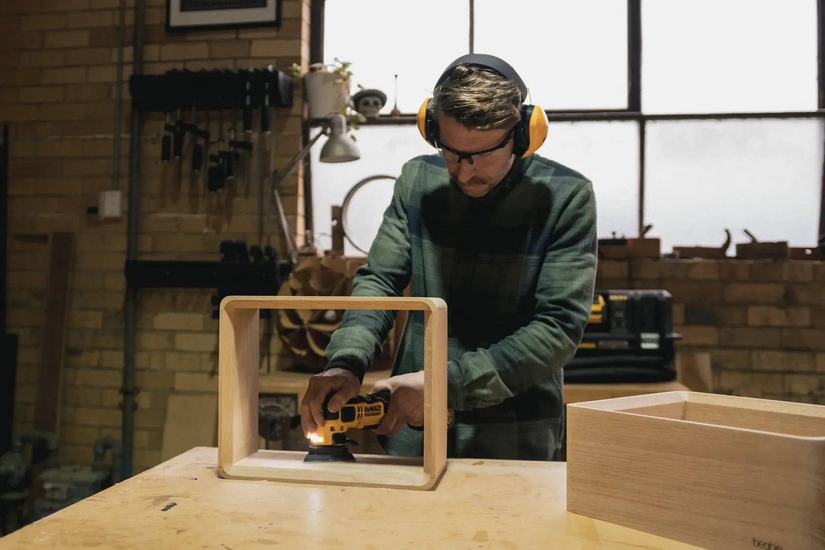 Persona lijando un marco de madera con lijadora DEWALT en un taller, usando protección auditiva y ocular.