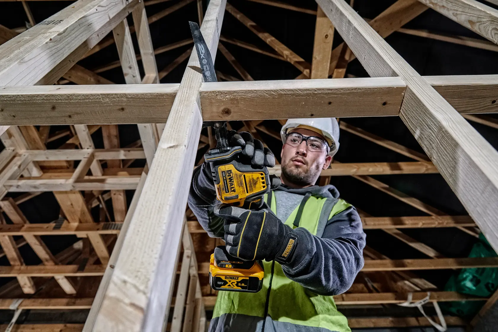 Un trabajador de la construcción con guantes y chaleco de seguridad usa una sierra recíproca inalámbrica DEWALT DCS369P2 para cortar una viga de madera en el marco de un edificio sin terminar.