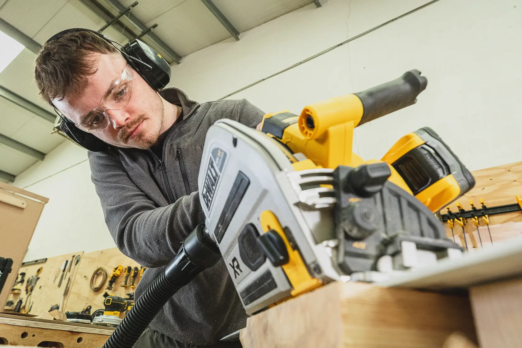Persona usando sierra circular inalámbrica DEWALT en madera, lleva gafas y protección auditiva en taller.