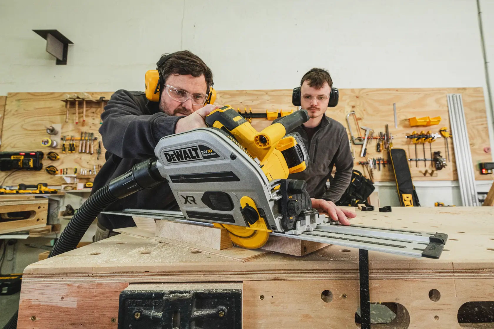 Dos personas usan una sierra de carril DEWALT inalámbrica para cortar madera en un taller.