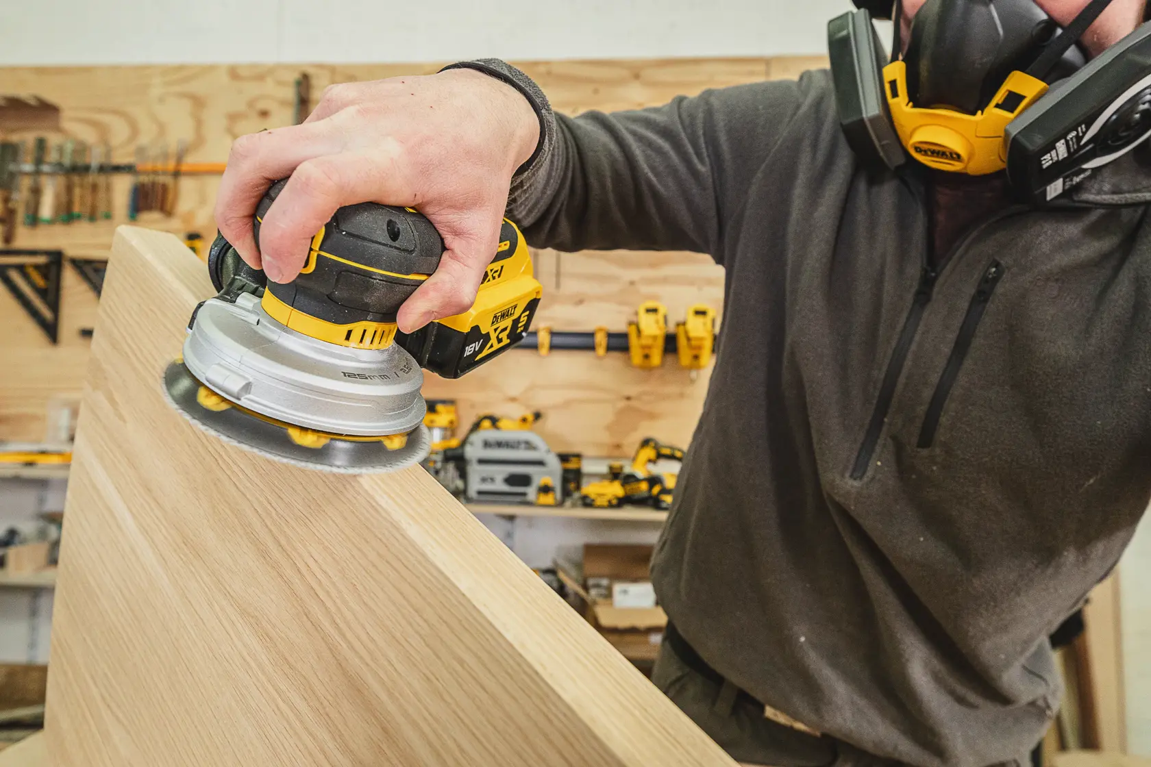 Persona lija madera con lijadora inalámbrica DEWALT en taller, usando mascarilla protectora.