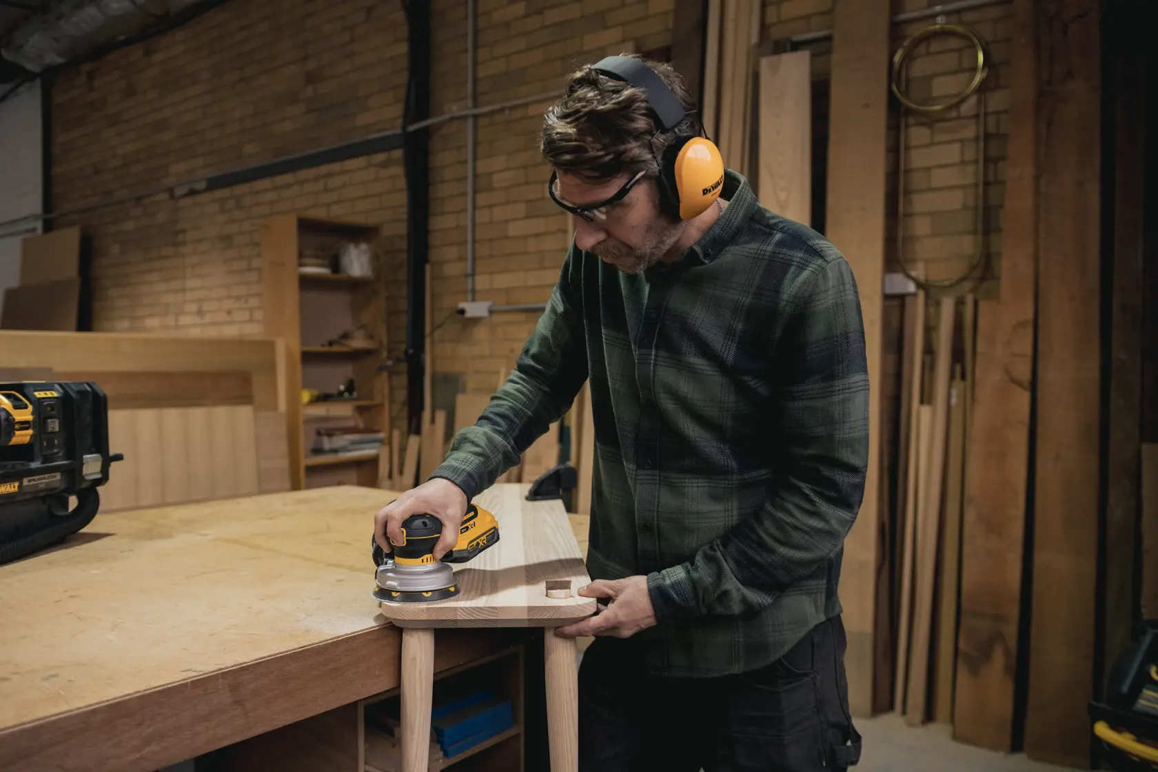 Persona lijando madera con lijadora inalámbrica DEWALT en taller, usando protección auditiva y ocular.