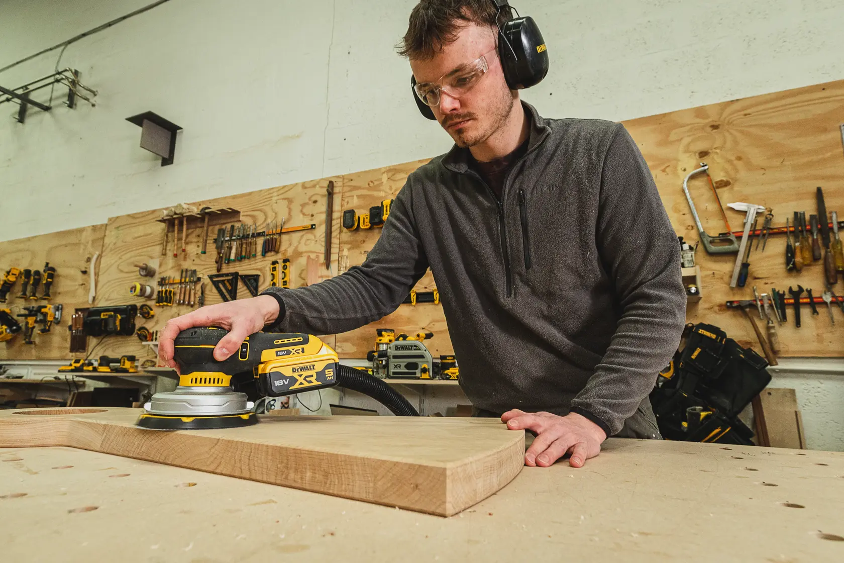 Persona lijando madera con una lijadora DEWALT DCW230N-XJ en taller, usando equipo de seguridad.