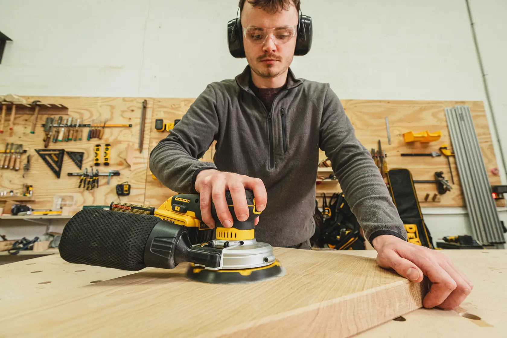 Persona usando una lijadora DEWALT sobre una tabla de madera en un taller con herramientas al fondo.
