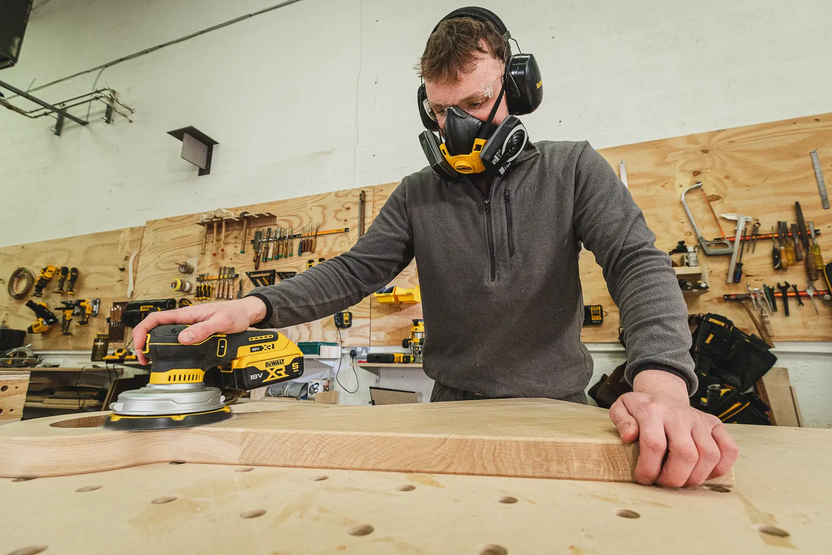 Persona lijando madera con una lijadora DEWALT, usando protección en taller con herramientas en la pared.