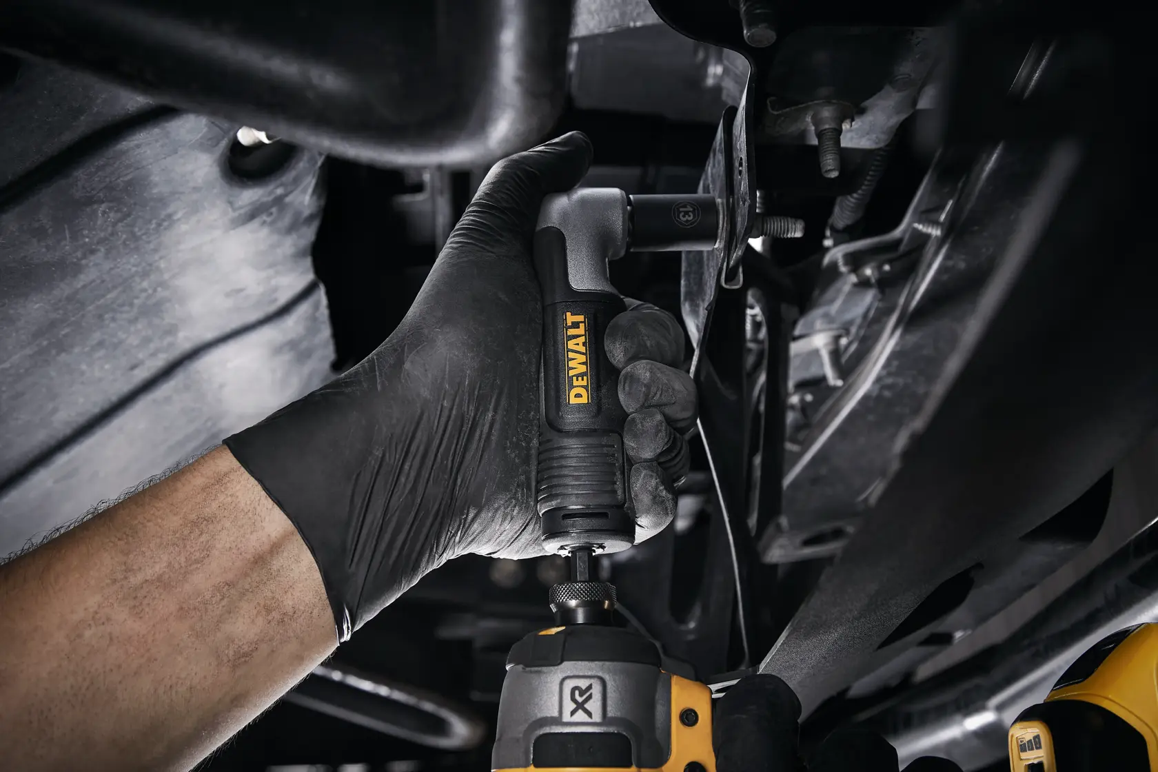 Mano con guante usa adaptador de taladro angular DEWALT bajo vehículo para espacios reducidos.