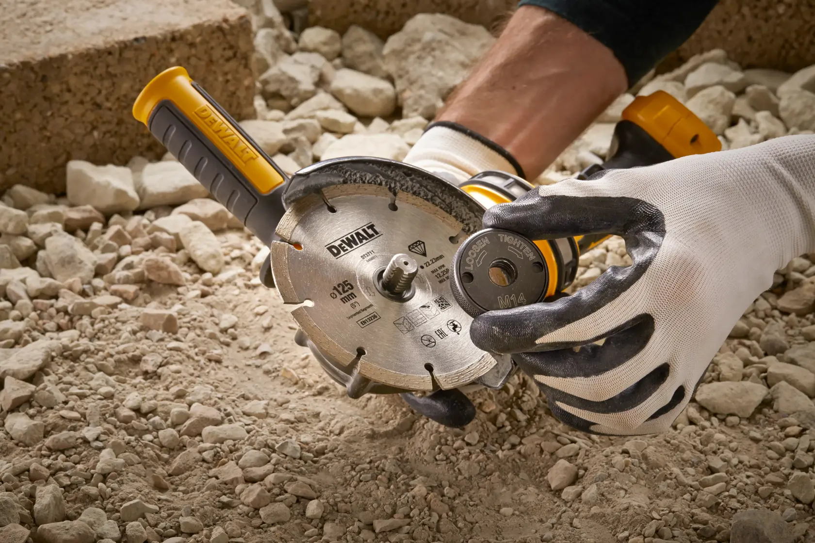 Manos con guantes sostienen una DEWALT DT3559 con disco de diamante sobre suelo pedregoso.