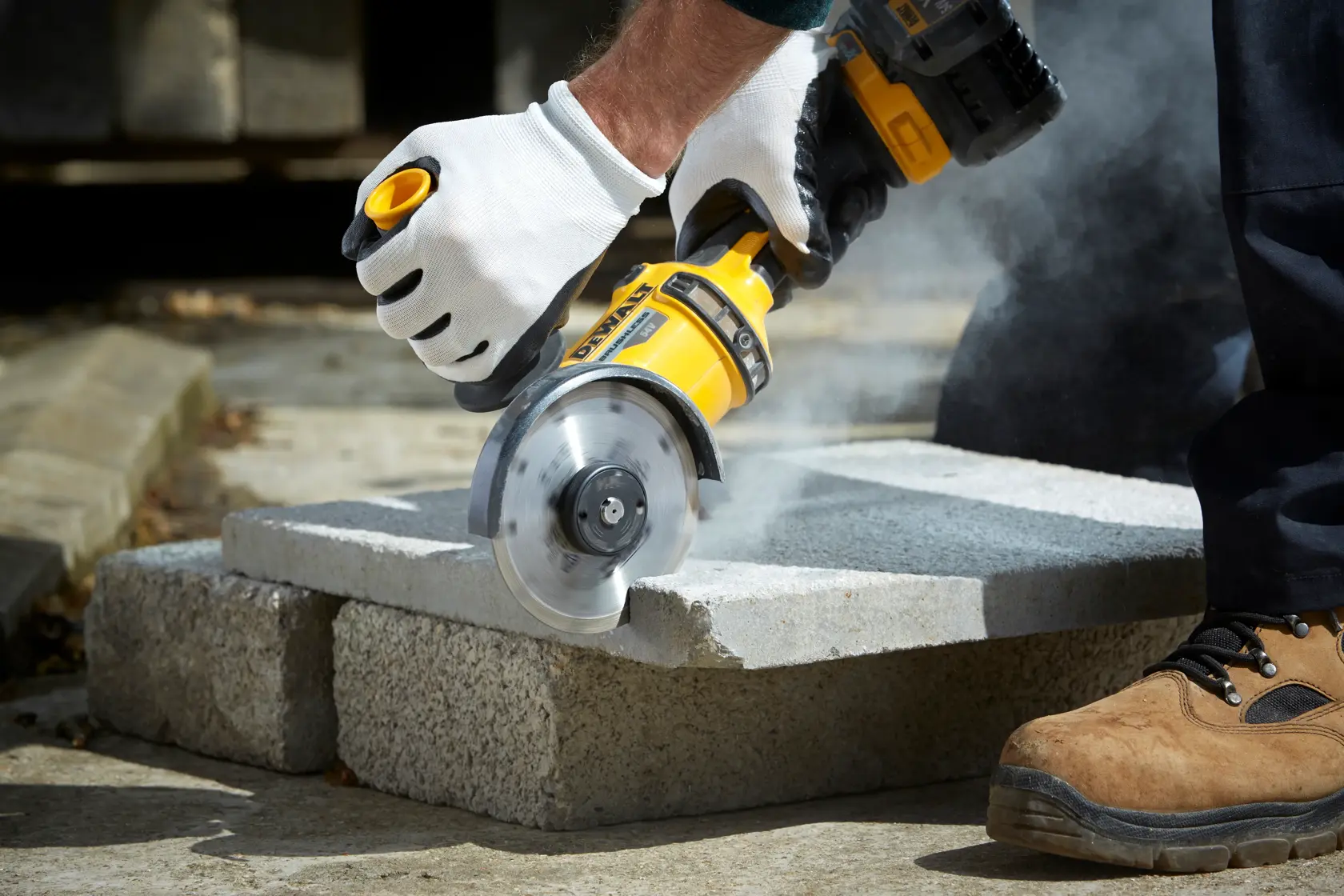 Persona corta bloque de cemento con esmeril DEWALT, con guantes y botas, polvo visible.