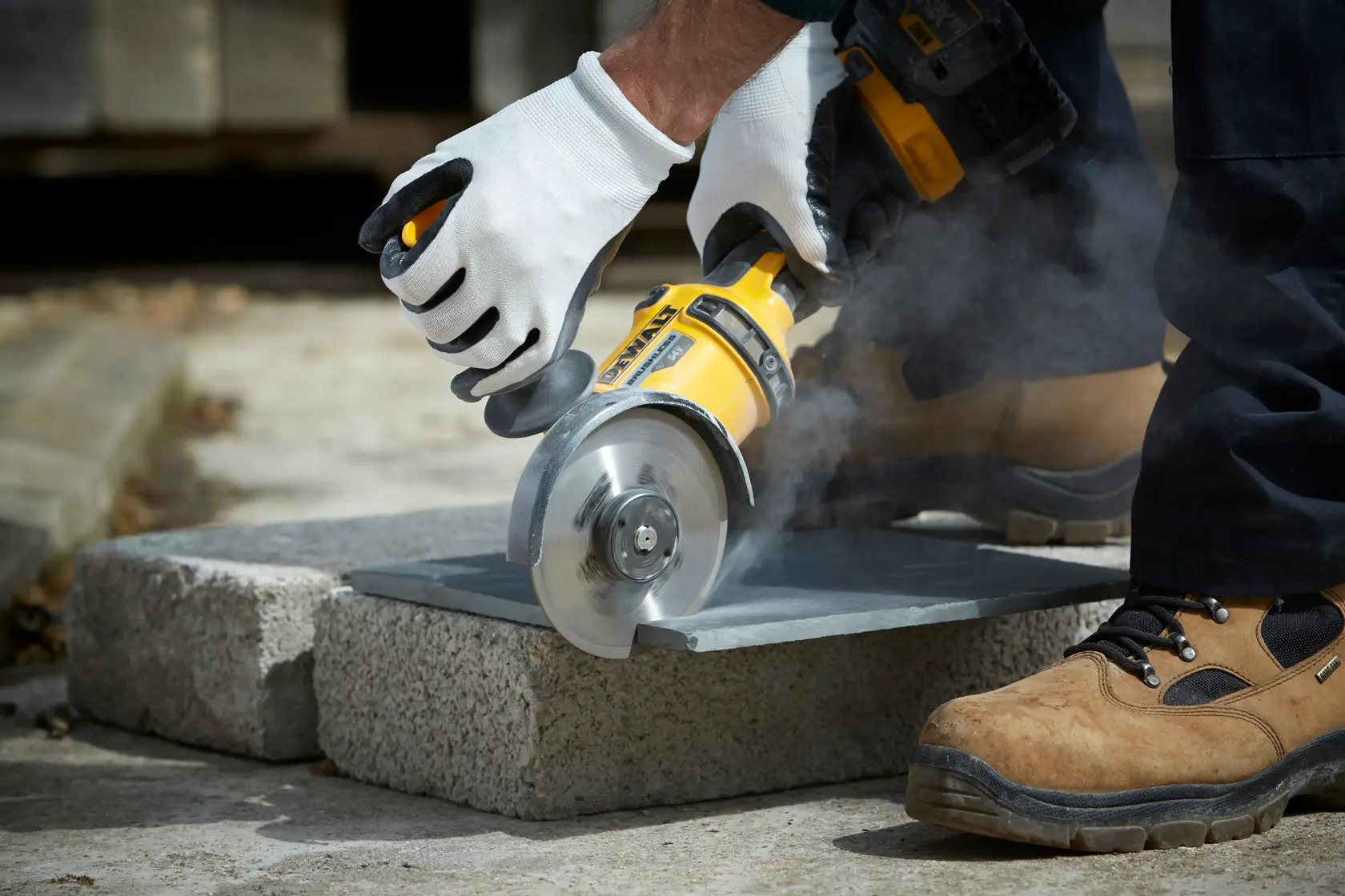 Manos con guantes usan una amoladora DEWALT para cortar piedra al aire libre, generando polvo.